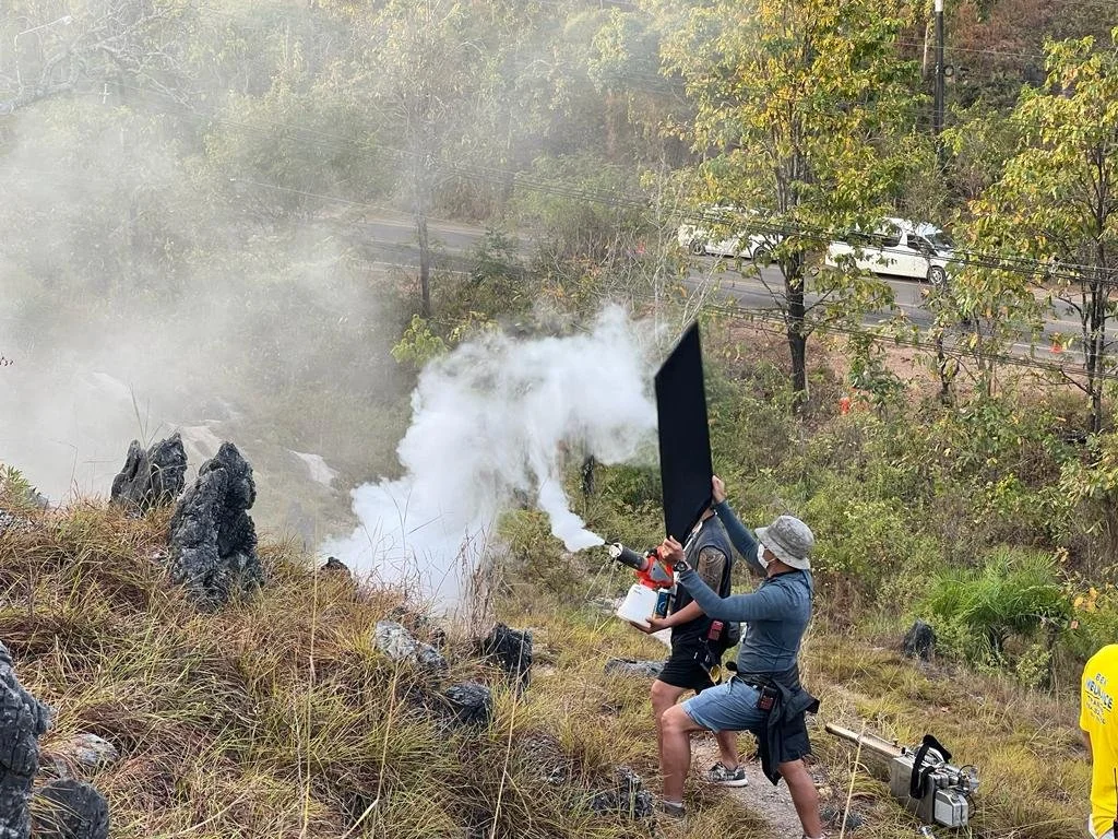 Two people are using a flame thrower to ignite a small fire on a rocky hillside with some tall grass, smoke, and trees in the background.