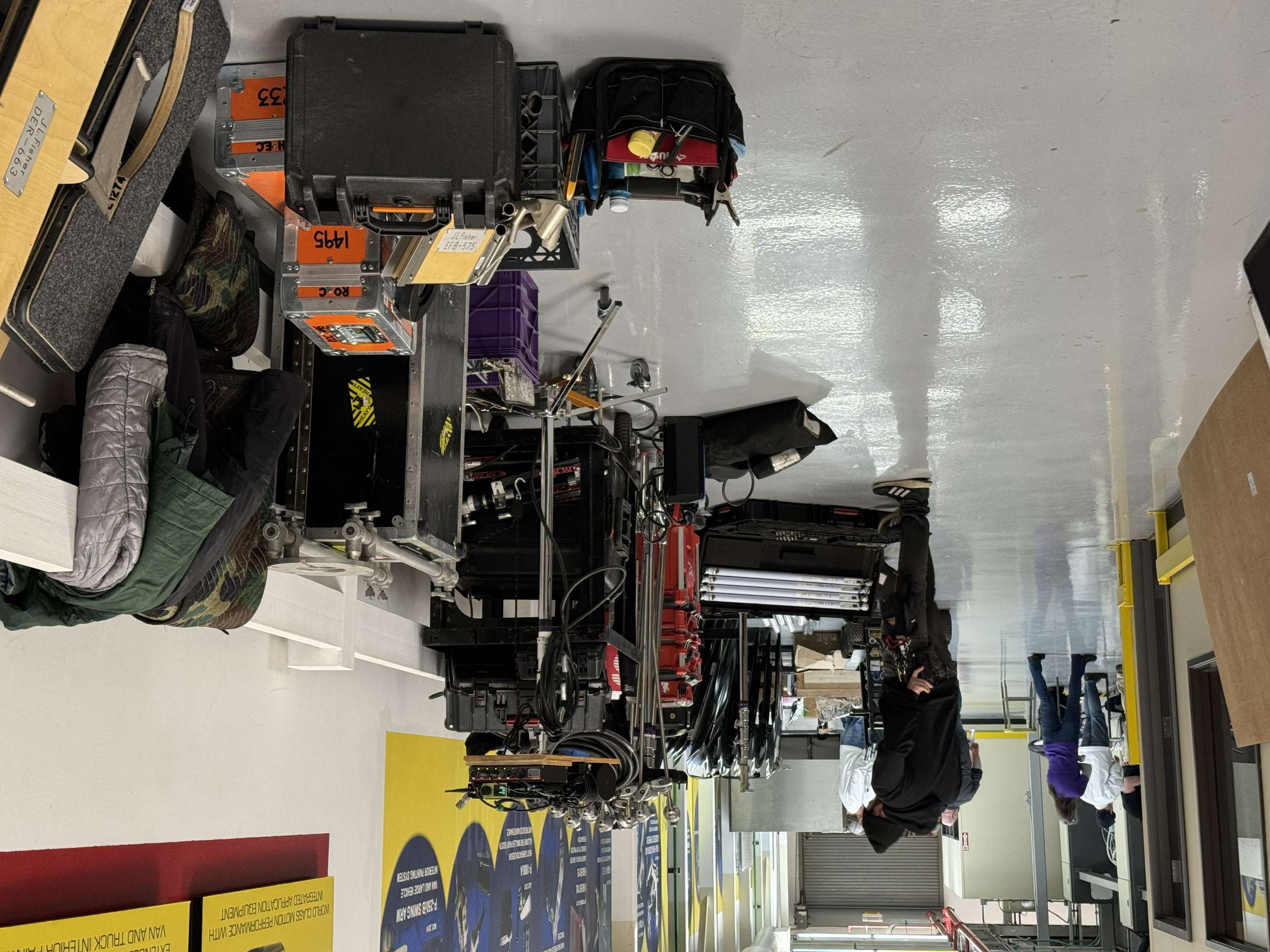 Workshop with various tools, equipment, and supplies, including black cases, colored bins, and workbenches. Several people are working or walking around the space.