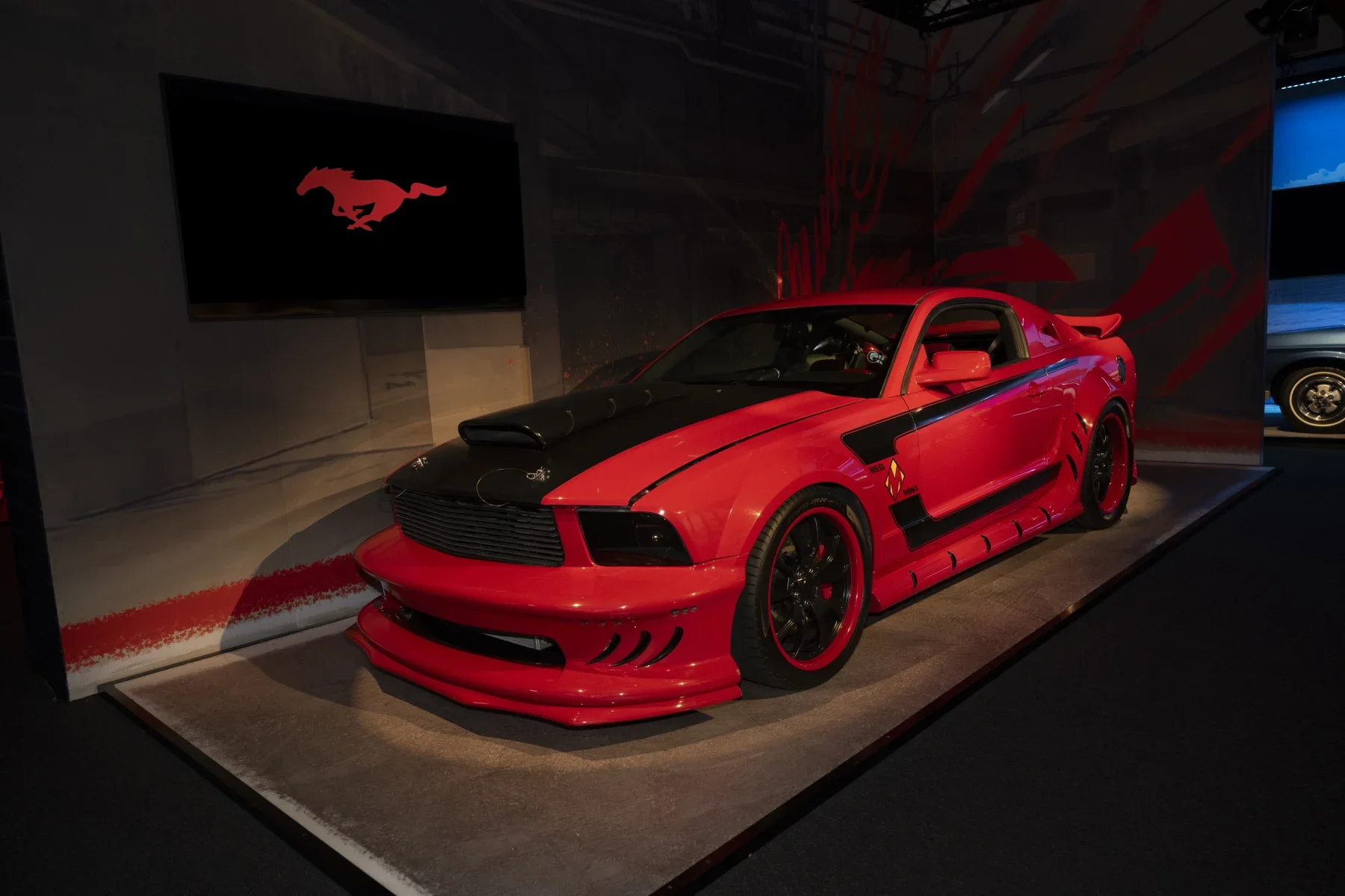 Red and black racing car on display at a museum, with Mustang logo on a screen behind it.