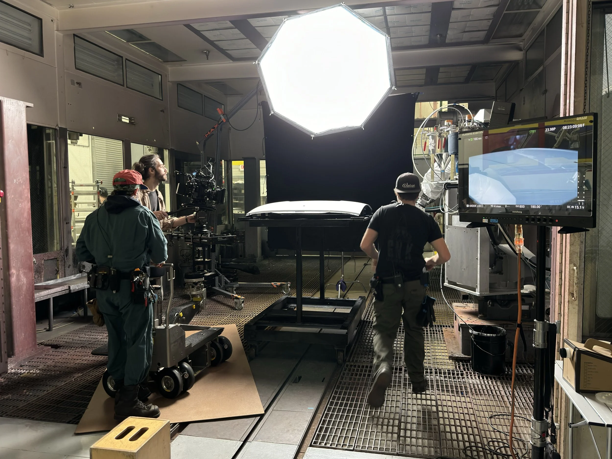 Film crew setting up camera and equipment inside the back of a truck with a black backdrop, large lighting umbrella overhead, and monitors for filming.