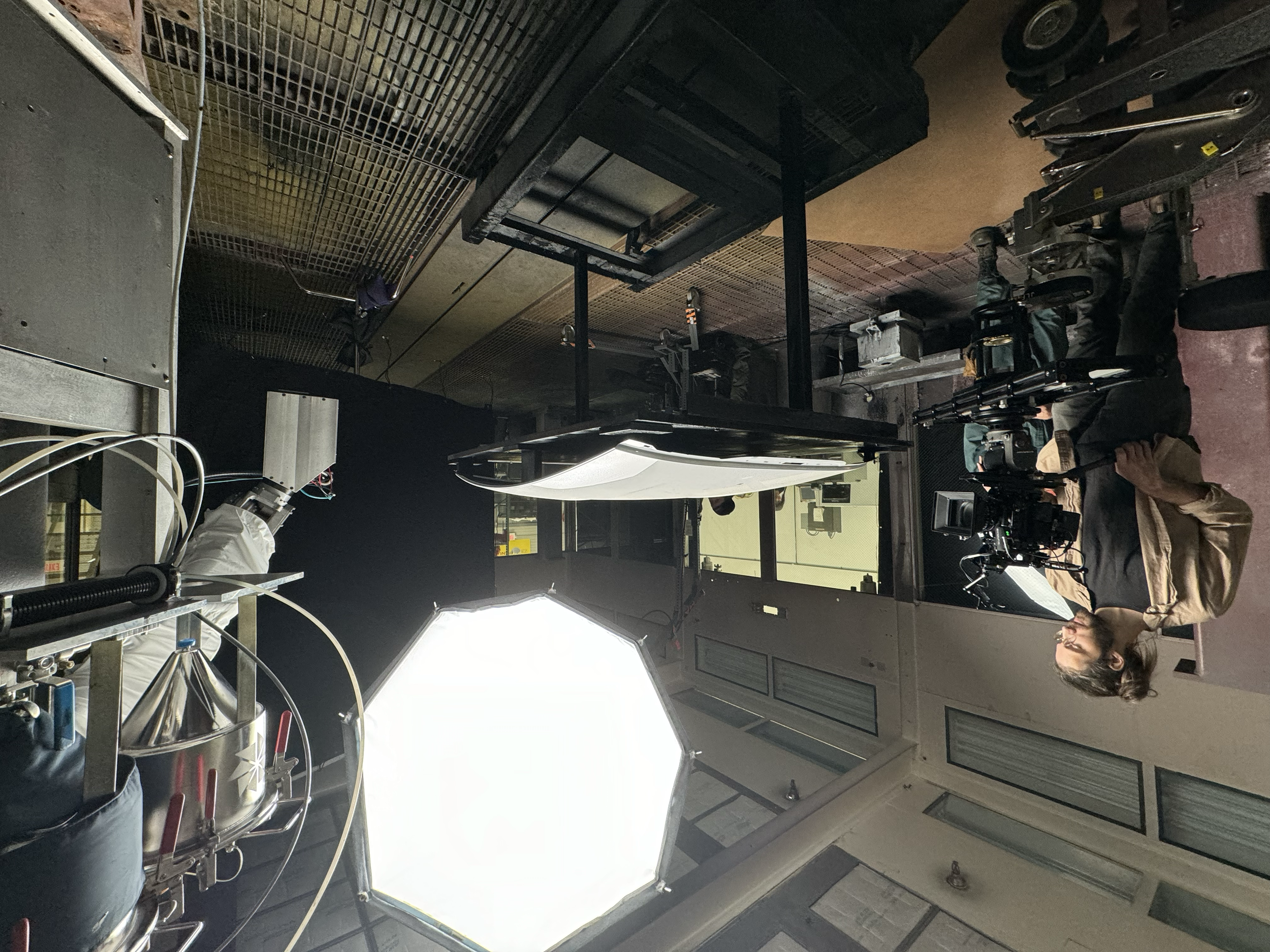 A woman with glasses and long hair sitting at a table with a camera rig, in a studio with professional lighting and equipment, with people working in the background.