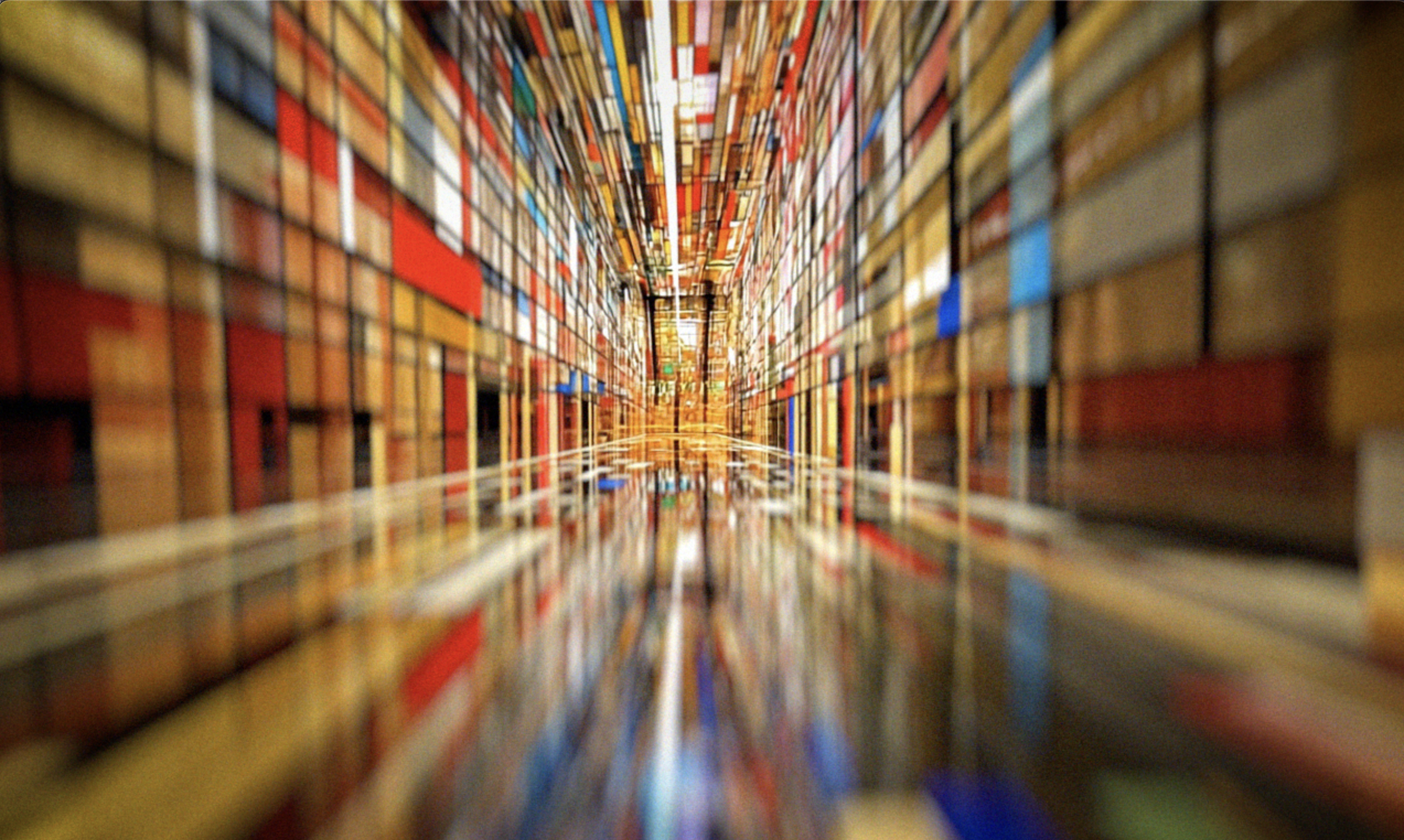 A low-angle view of a long library aisle filled with colorful bookshelves, with books stacked vertically on both sides and reflected on the polished wooden floor.