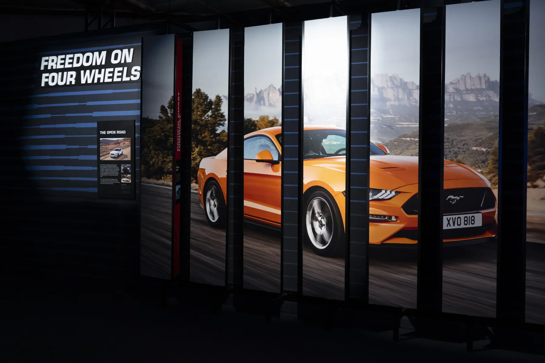 An orange Ford Mustang car displayed on a large vertical digital screen with the slogan 'FREEDOM ON FOUR WHEELS' and a smaller text panel titled 'THE OPEN ROAD' in the background.