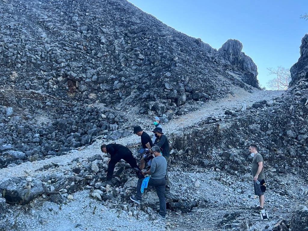 Group of people, some wearing masks, assisting each other while climbing a rocky slope in a mountainous area on a clear day.