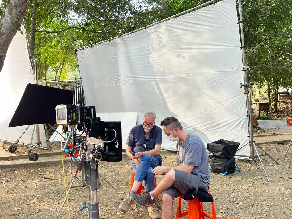 Two men wearing face masks sit on orange stools in an outdoor film production set, surrounded by a white backdrop, lighting equipment, and trees.