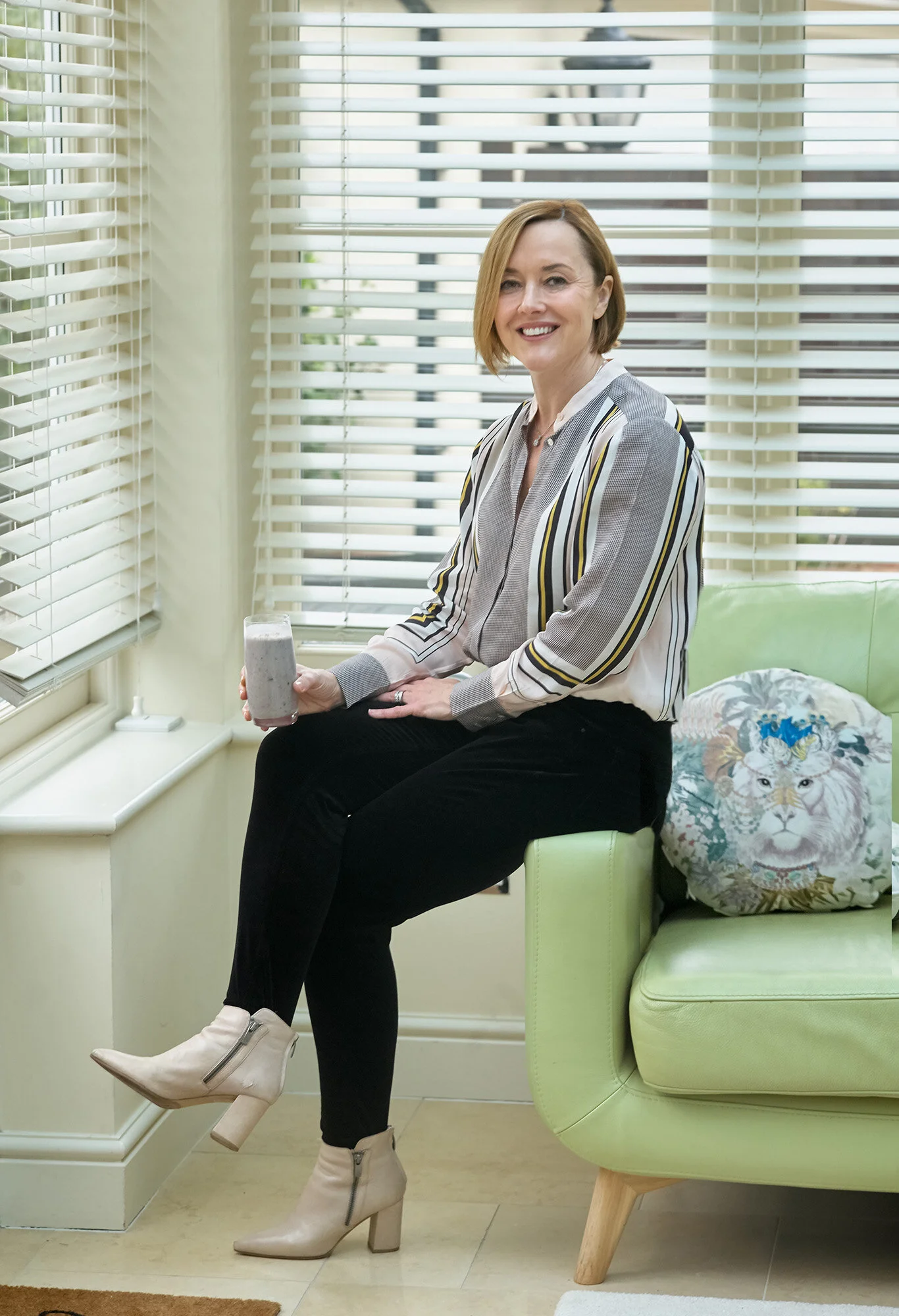 A woman with short, light brown hair smiling while sitting on a light green sofa near windows with horizontal blinds, holding a tall glass of a smoothie or milkshake, wearing a striped blouse, black pants, and beige ankle boots with heels.