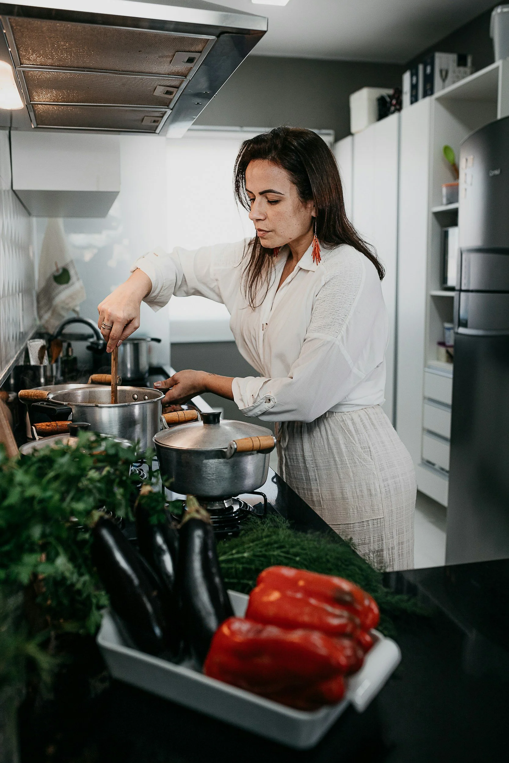 A woman cooking in a modern kitchen with fresh vegetables including eggplants, red bell peppers, and herbs on the counter.