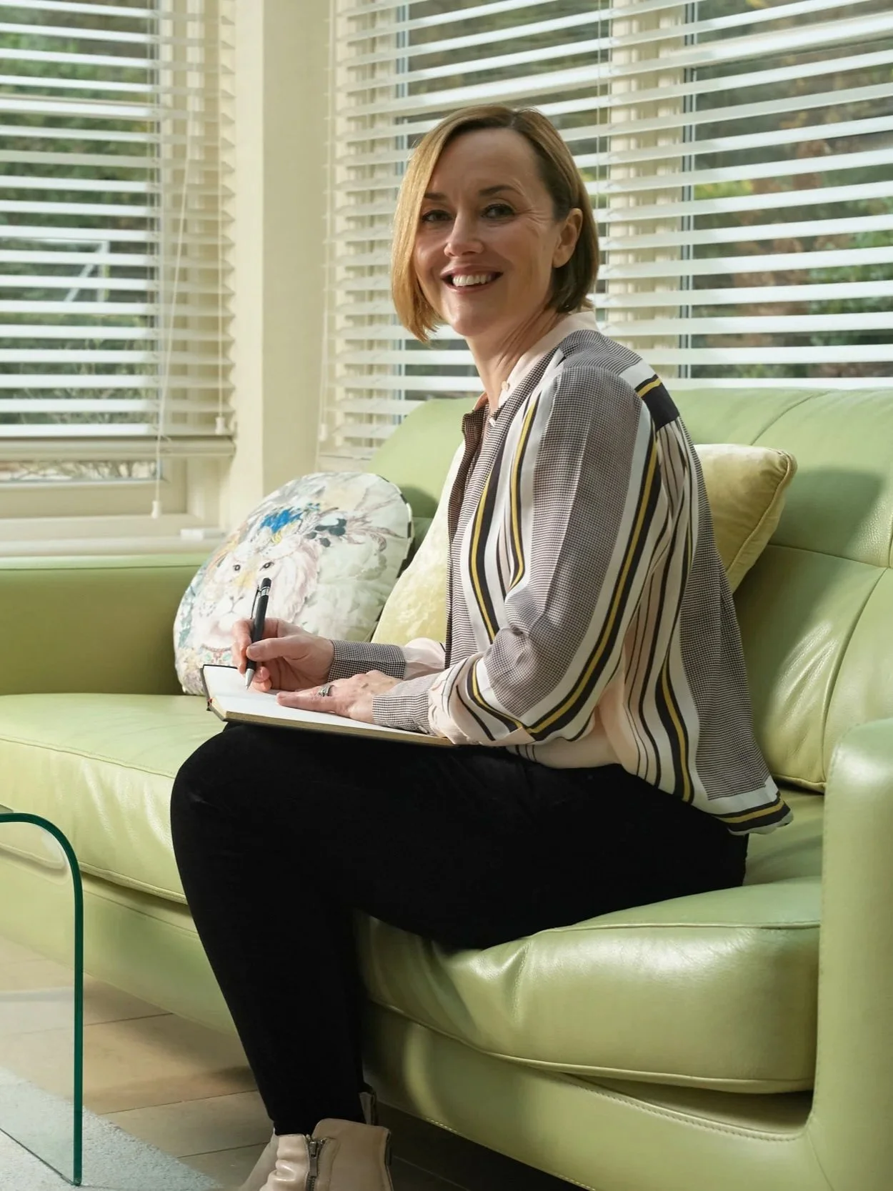 A woman with short brown hair smiling while sitting on a lime green sofa, holding a notebook and pen, near a window with white blinds.