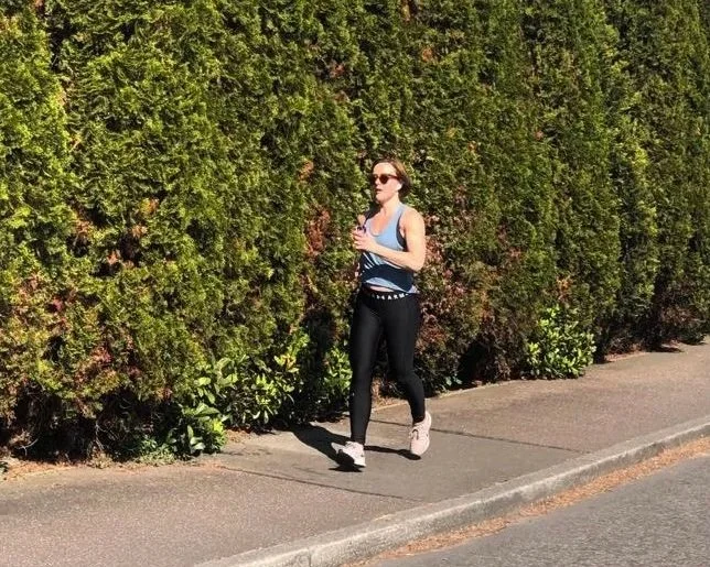 A woman jogging on a pavement next to a tall green hedge, wearing sunglasses, a blue tank top, black leggings, and white running shoes.