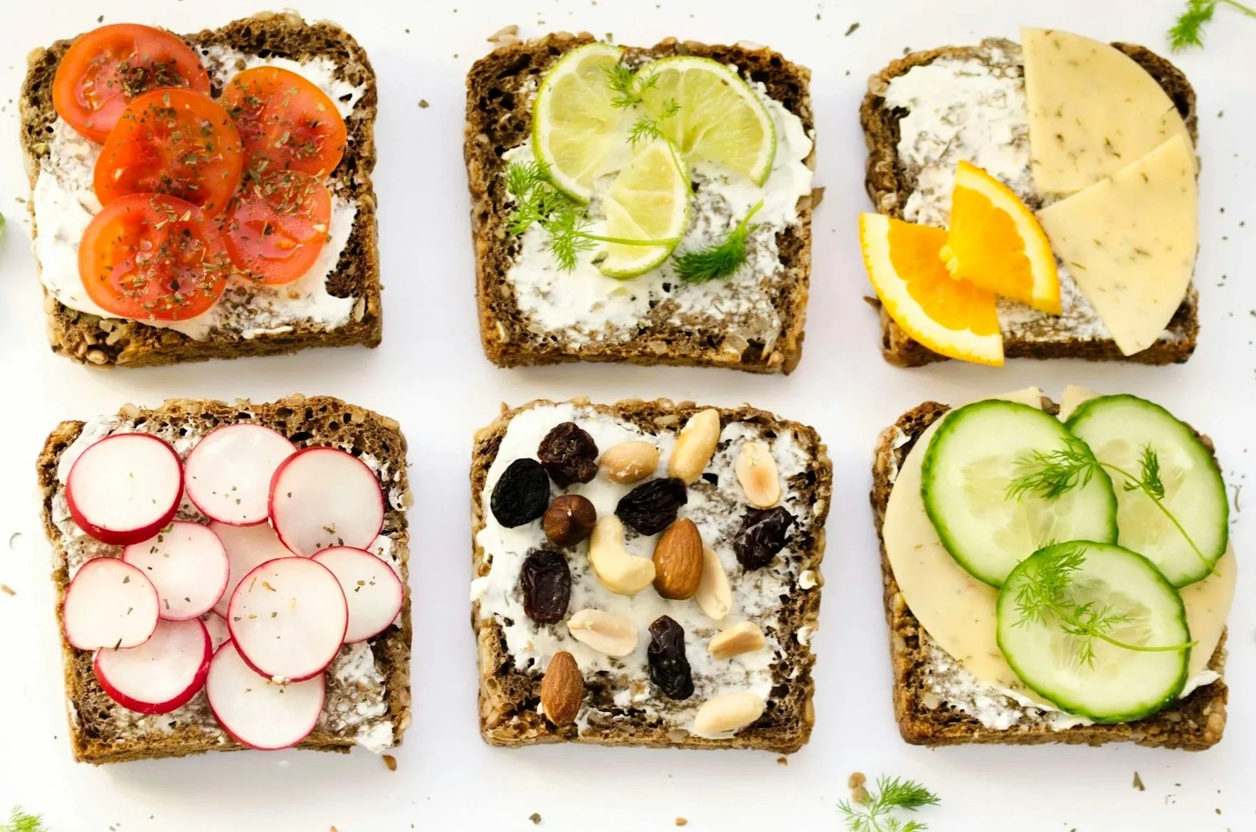 Six slices of toast with various toppings arranged in two rows of three on a white surface. Top row: cherry tomato with herbs, lemon slices with herbs, cheese with orange pieces. Bottom row: radishes with herbs, mixed nuts and dried fruit with cream, cucumber slices with herbs.