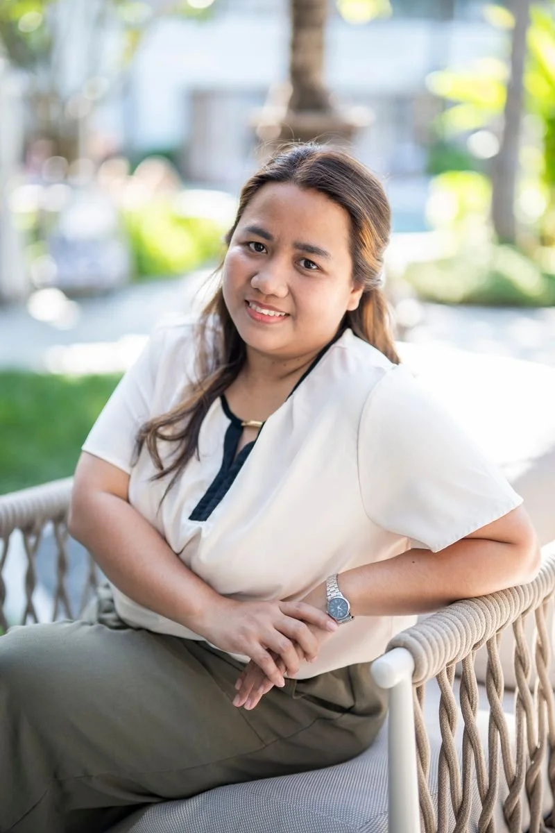 A woman in a cream-colored blouse and dark blue jeans standing outdoors on a pathway in a park with greenery and trees in the background.