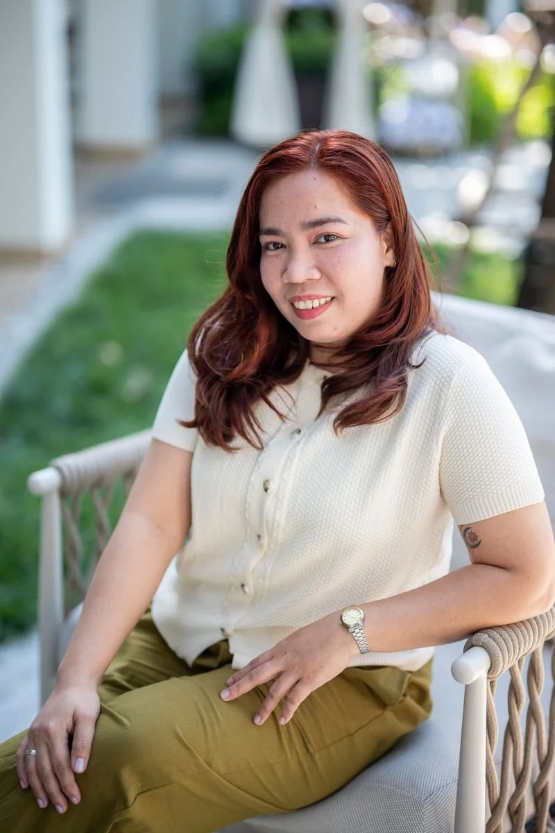 A woman in a white t-shirt and blue jeans standing outdoors on a curved pathway with green trees and grass in the background.