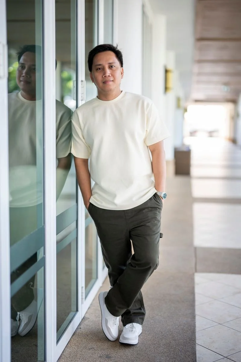 A man with short black hair and a white shirt standing outdoors on a curved pathway with green grass and palm trees, arms crossed, smiling at the camera.