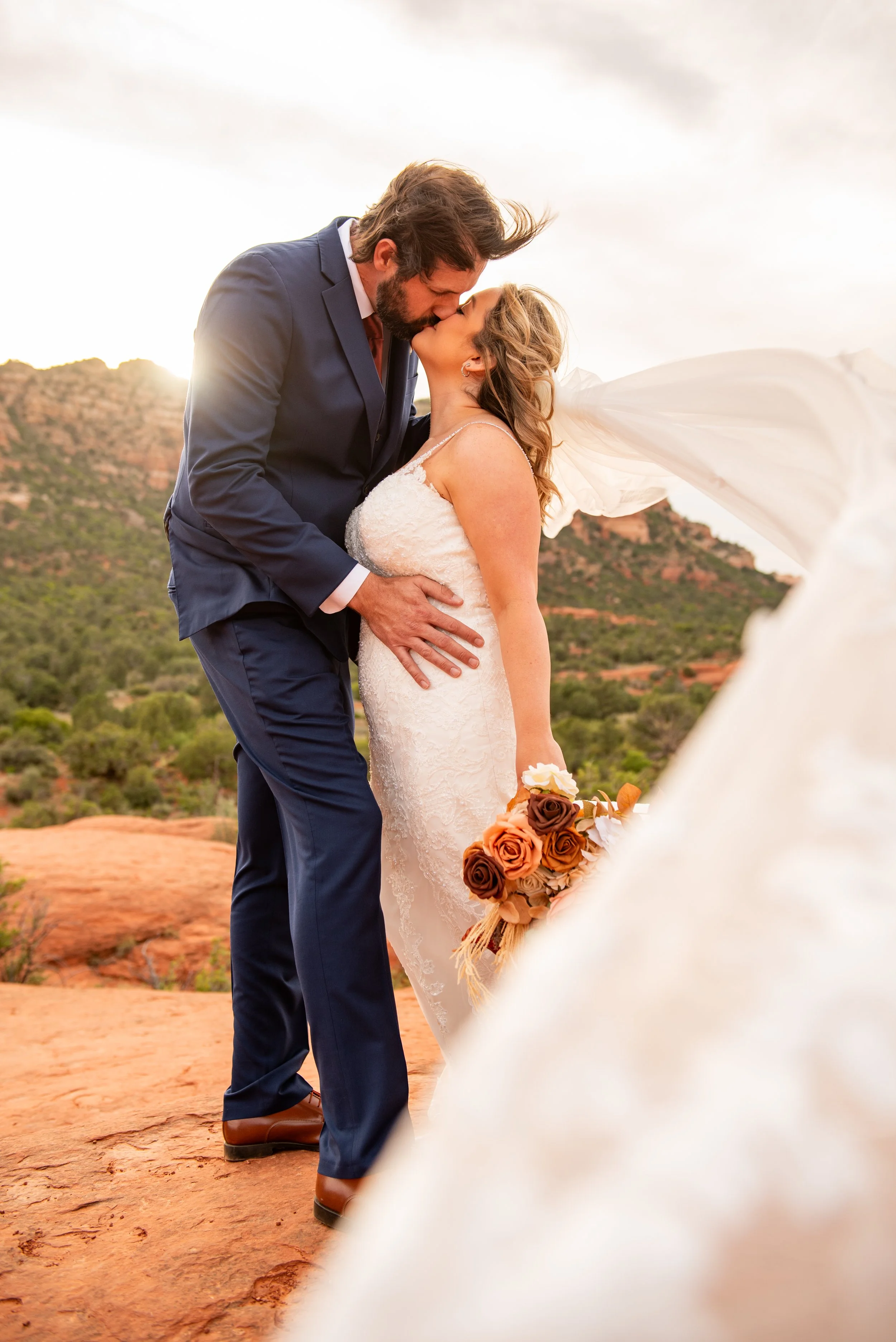 The bride and groom share a kiss while the brides cathedral veil wraps around them while surrounded by the red rocks of Sedona.