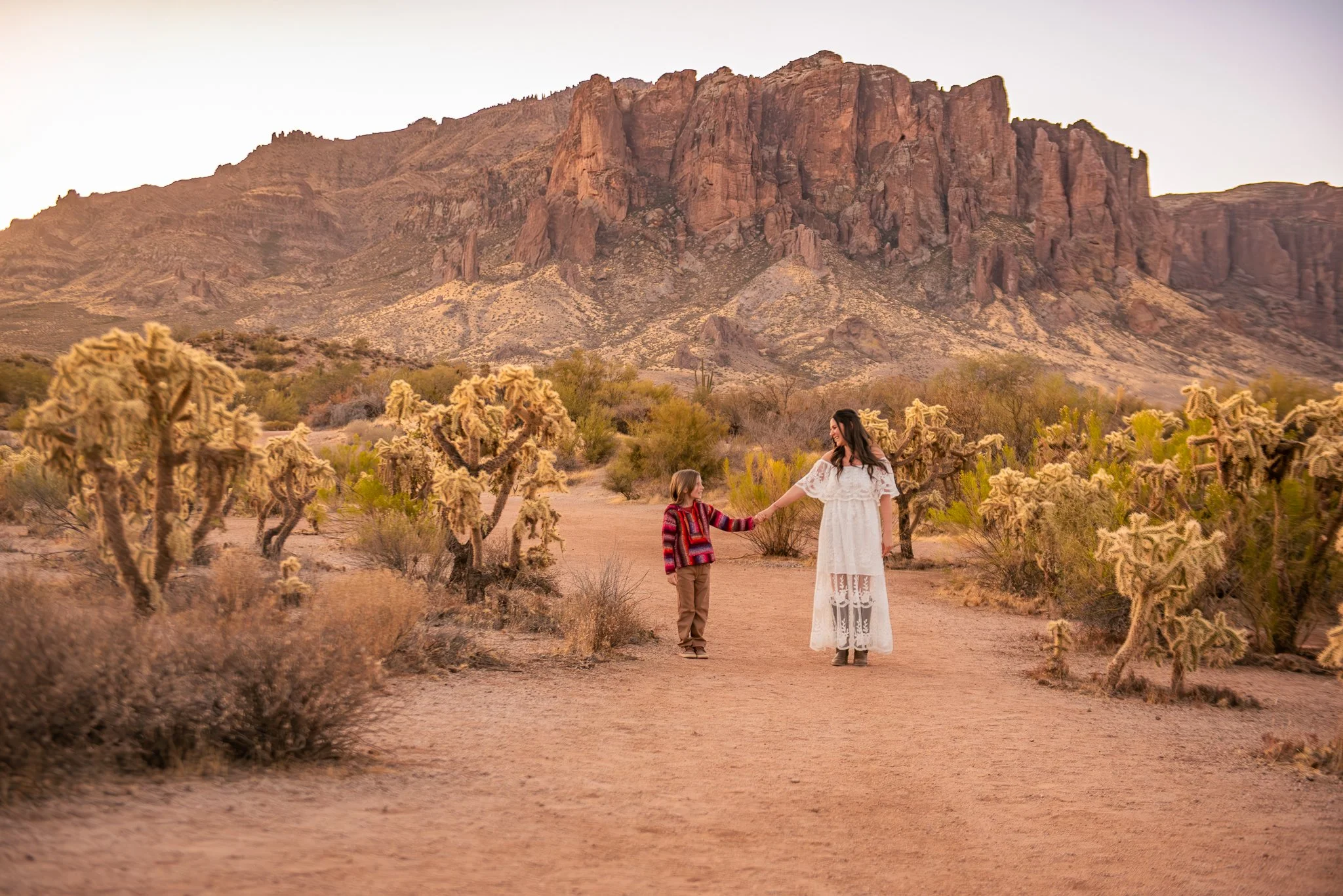 Mother and son hold hands and look at one another at this sunrise family session with the majestic superstition mountains standing tall behind them at First Water Trailhead in Apache Junction