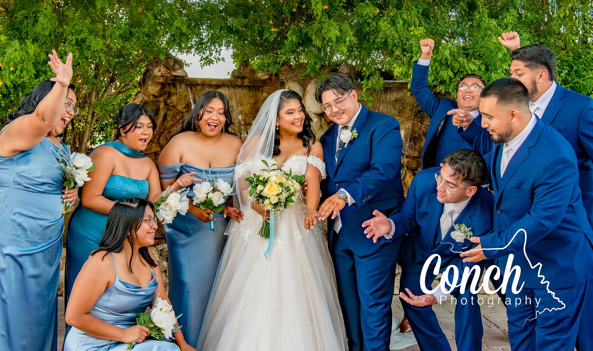 The wedding party gathers around the bride and groom cheering as the happy couple show off their wedding rings in this vibrant, true to color wedding photo.