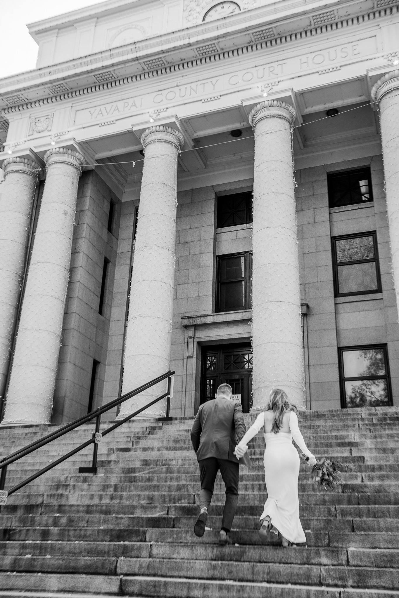  Classic black and white image featuring the eloping bride and groom running up the courthouse stairs holding hands during their destination adventure elopement at Downtown Prescott in Prescott, Arizona. 