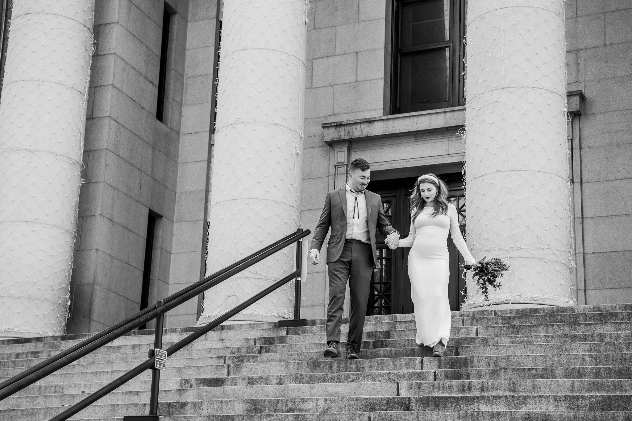  Classic black and white image featuring the eloping bride and groom running down the courthouse stairs holding hands during their destination adventure elopement at Downtown Prescott in Prescott, Arizona. 