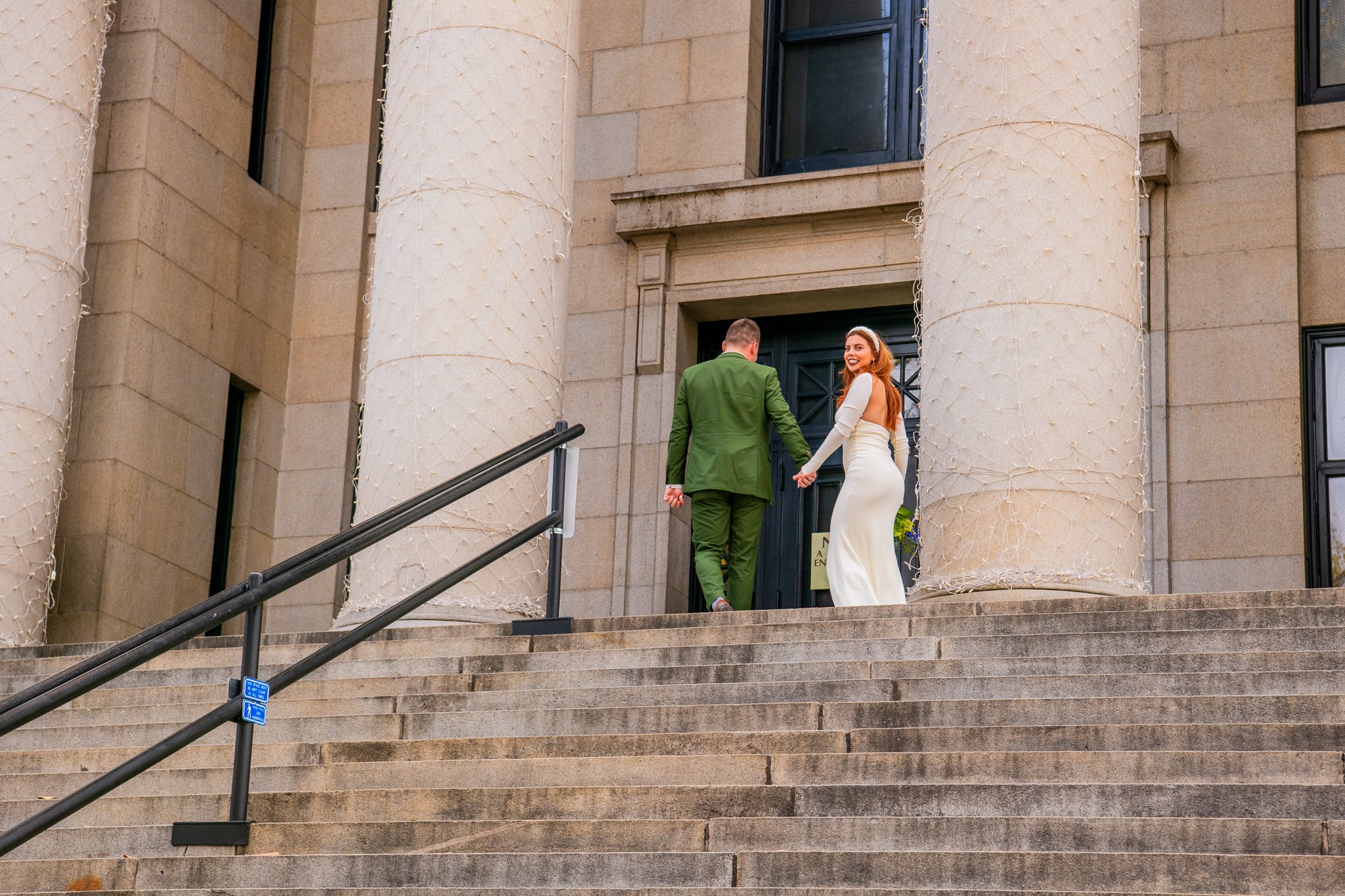  Bride and groom standing at the top of the courthouse steps, bride peaking behind her shoulder at the Yavapai Courthouse in Downtown Prescott, Arizona during their destination adventure elopement. 
