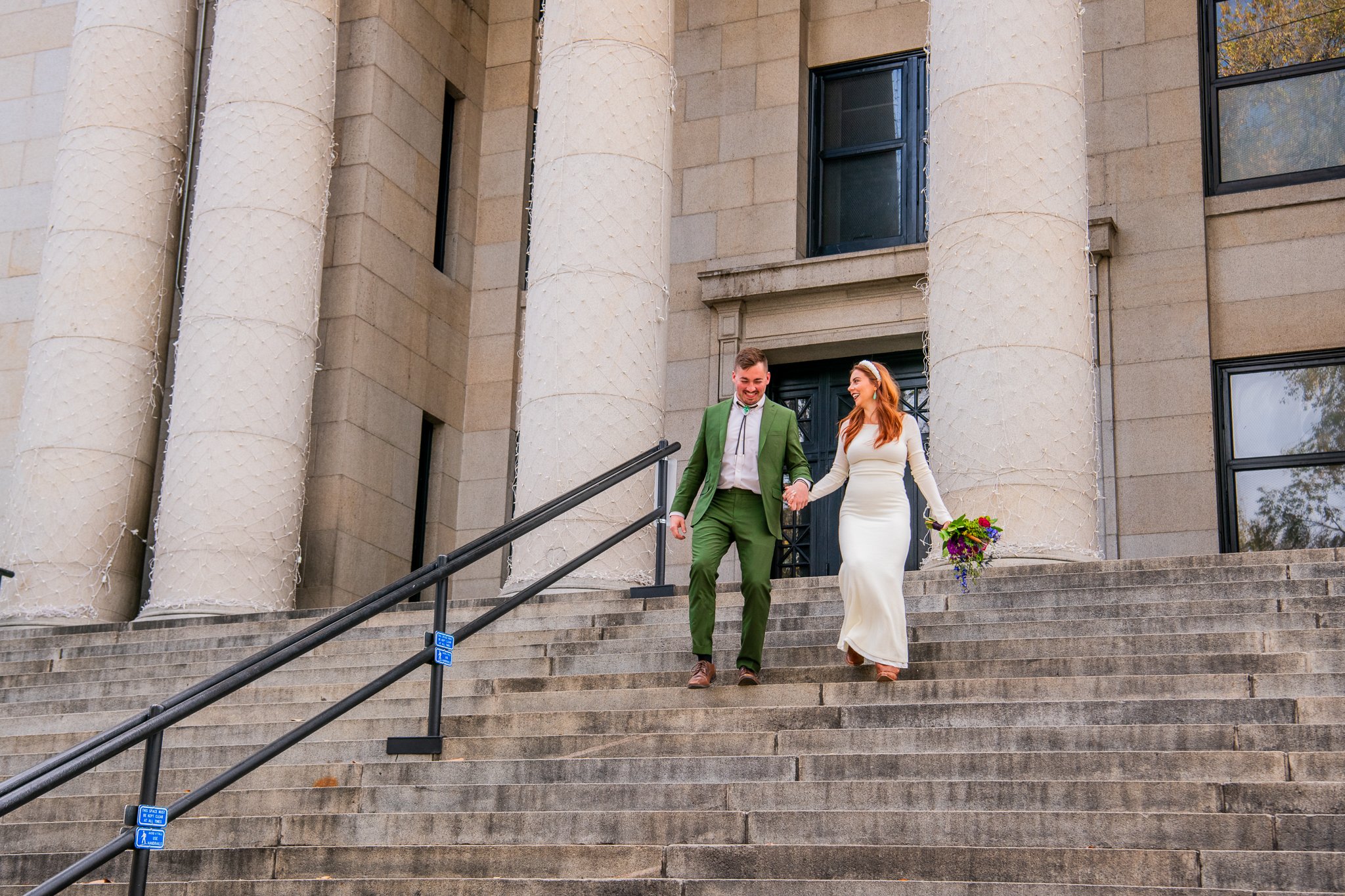  Classic, timeless image featuring the eloping bride and groom running down the courthouse stairs holding hands during their destination adventure elopement at Downtown Prescott in Prescott, Arizona. 