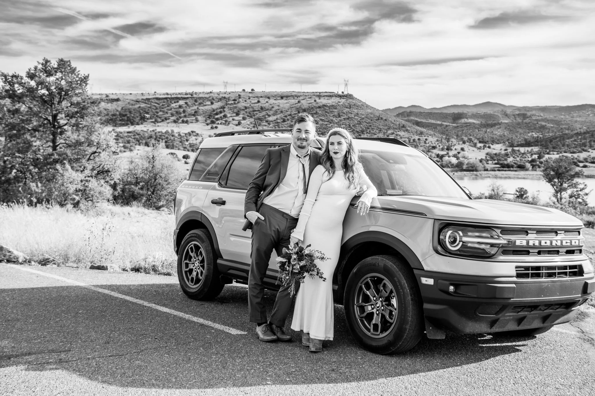 Black and white image of bride and groom stand against their rented Ford Bronco during their destination adventure elopement at Watson Lake in Prescott, Arizona.