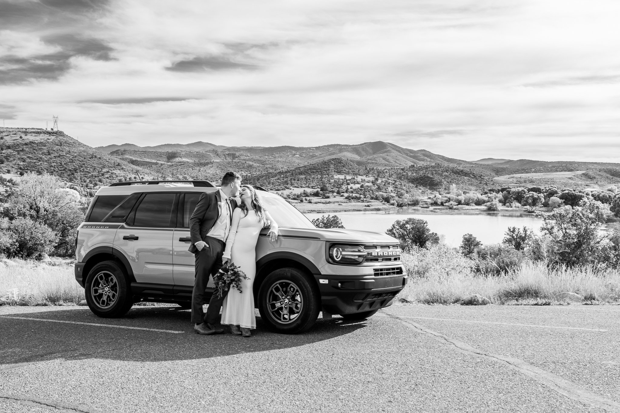 Black and white landscape image of bride and groom kissing while leaning on their rented Bronco at Watson Lake in Prescott, Arizona during their Destination Adventure Elopement.