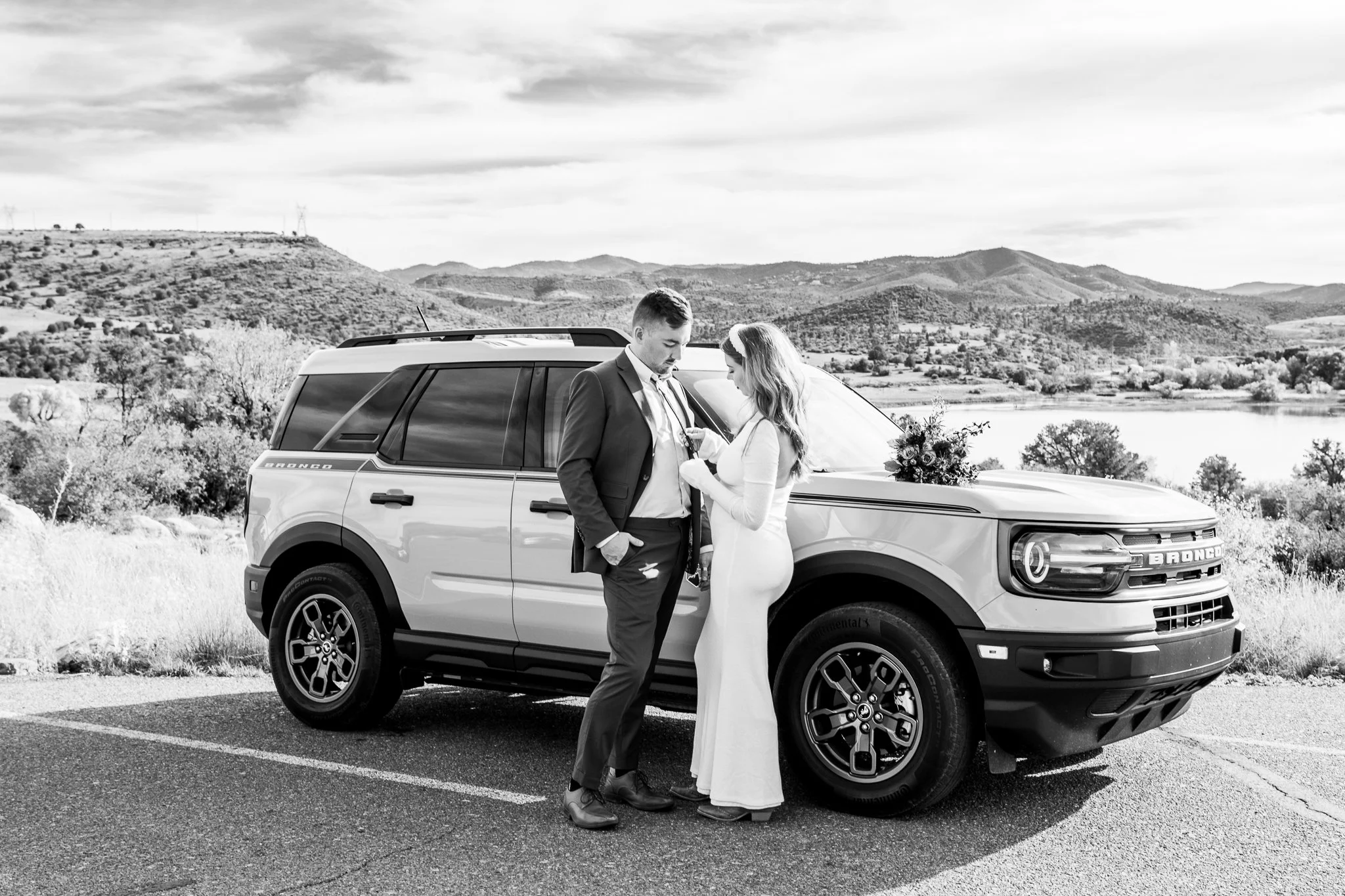 Black and white image of bride fixing groom's tie in front of their rented Ford Bronco at Watson Lake in Prescott, Arizona during their destination adventure elopement.