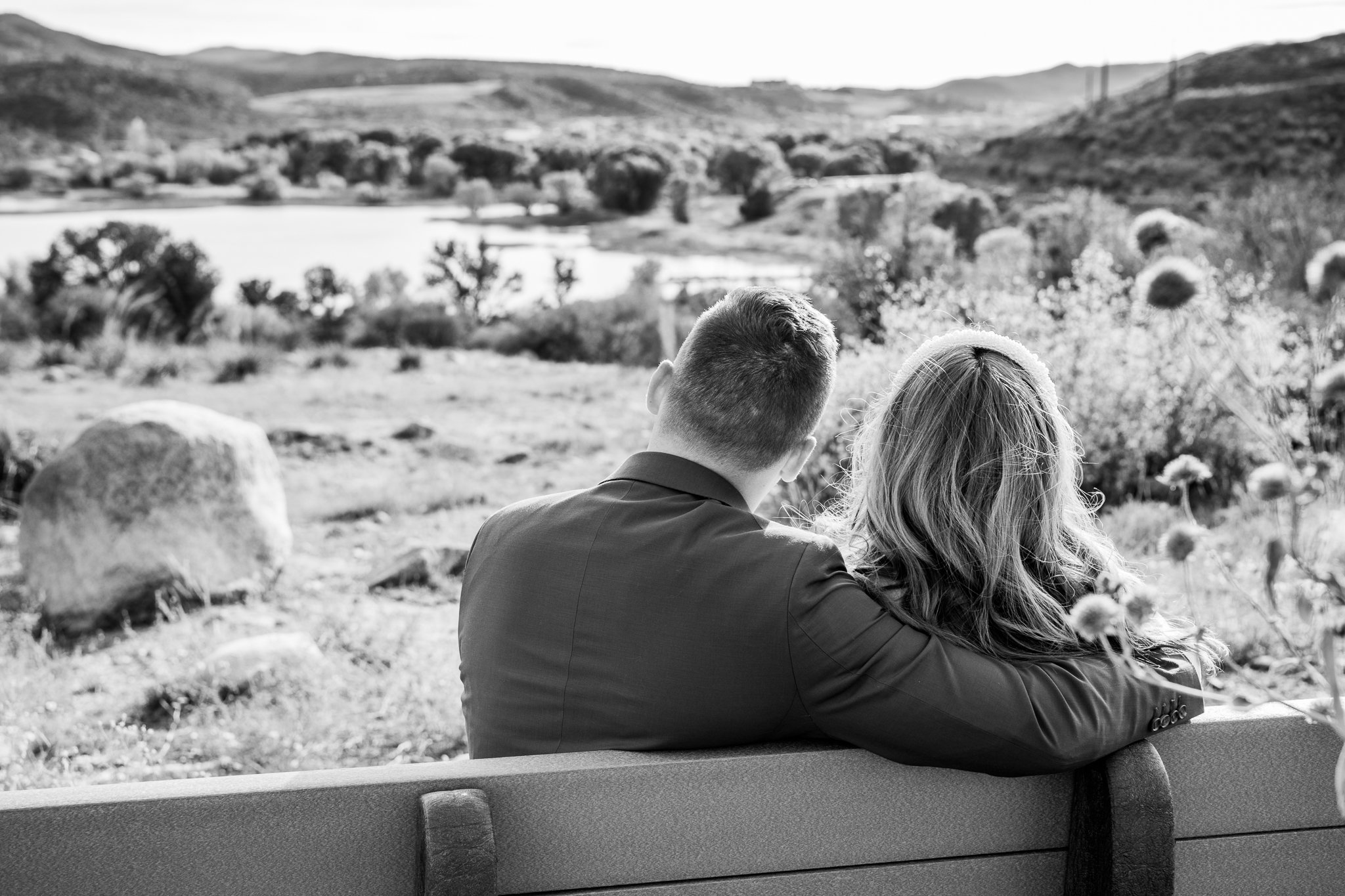 Sentimental black and white image of the backside of the bride and groom sitting each other on a bench at Watson Lake in Prescott, Arizona during their destination adventure elopement.