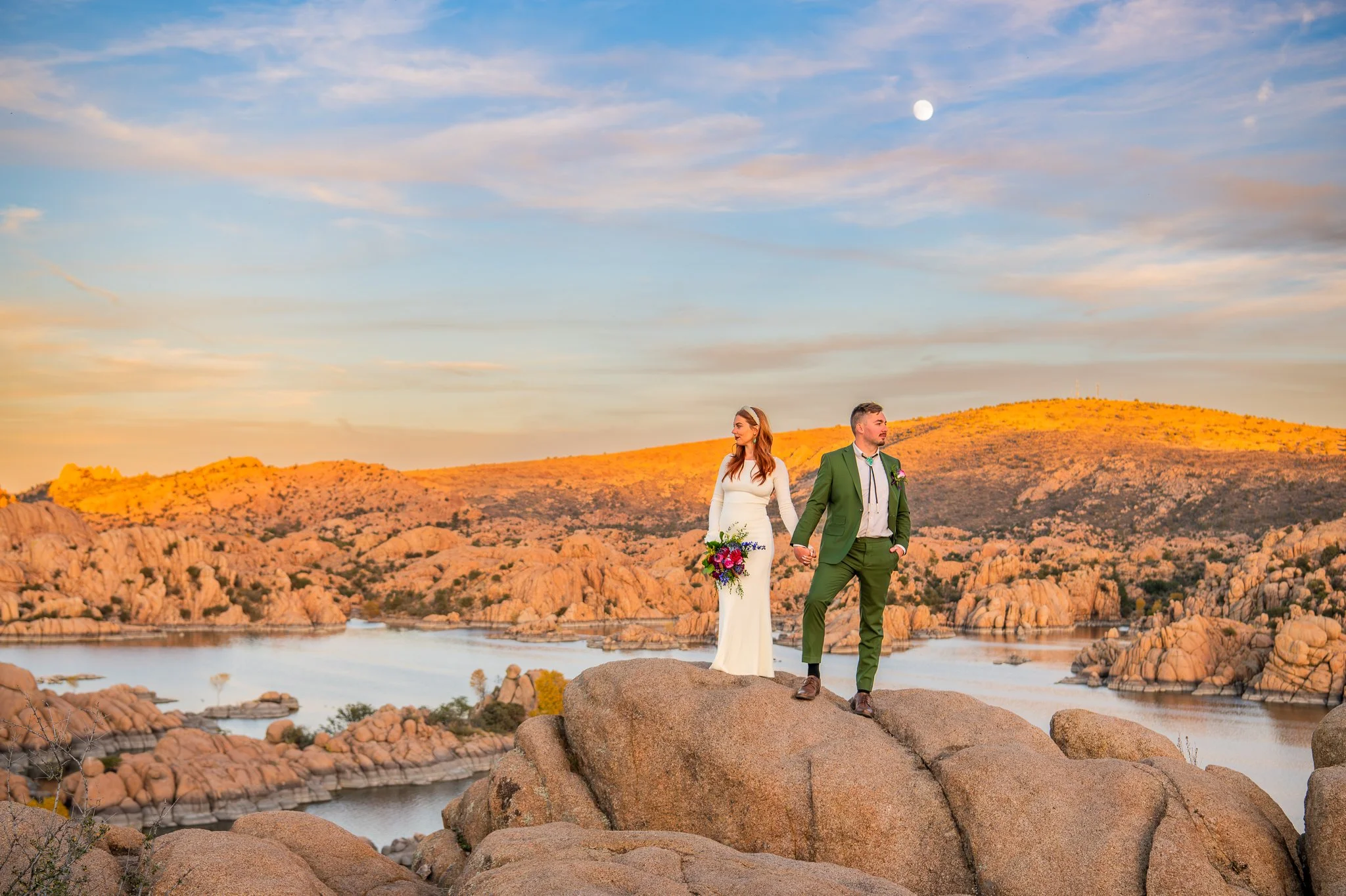 Bride and Groom hold hands while standing on the large granite rocks at Watson Lake in Prescott, Arizona looking at the sun kissed land around them while the full moon rises at sunset.