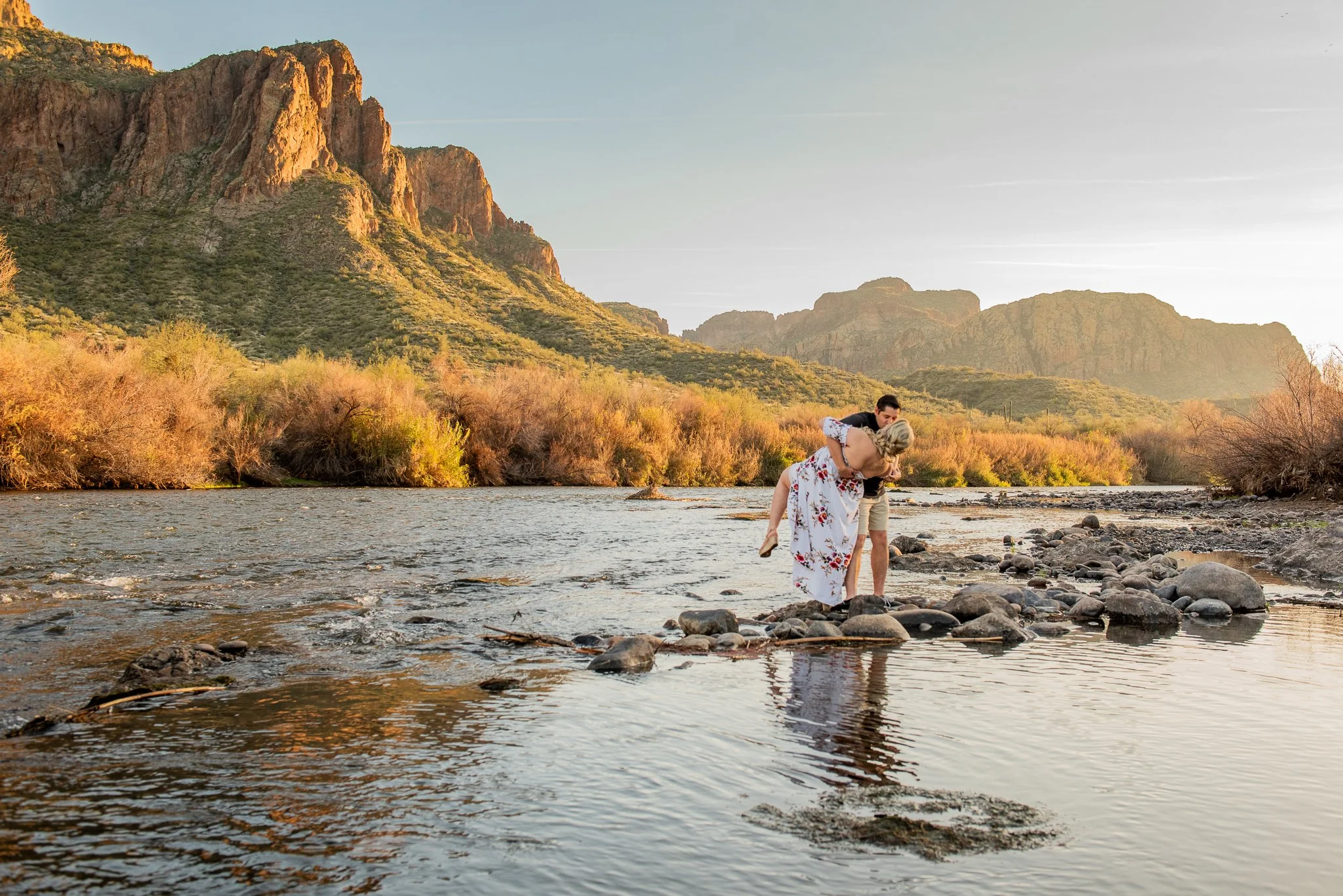 A women gets a dip kiss from her fiance at sunset in the Salt River in Mesa, Arizona with Mountains surrounding them during golden hour.