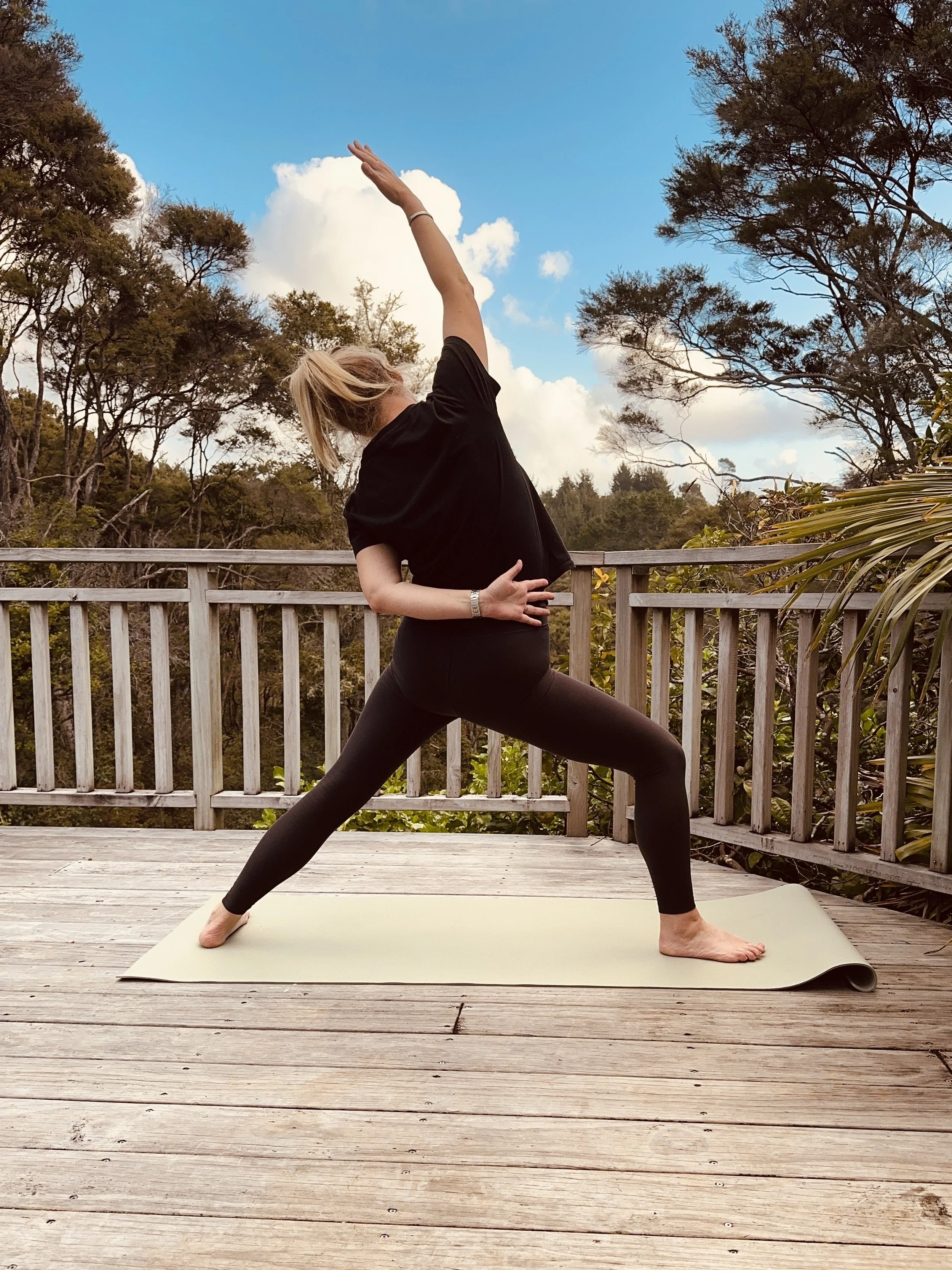 A woman practicing yoga outdoors on a wooden deck, performing a side stretch pose on a yoga mat against a backdrop of trees and a partly cloudy sky.