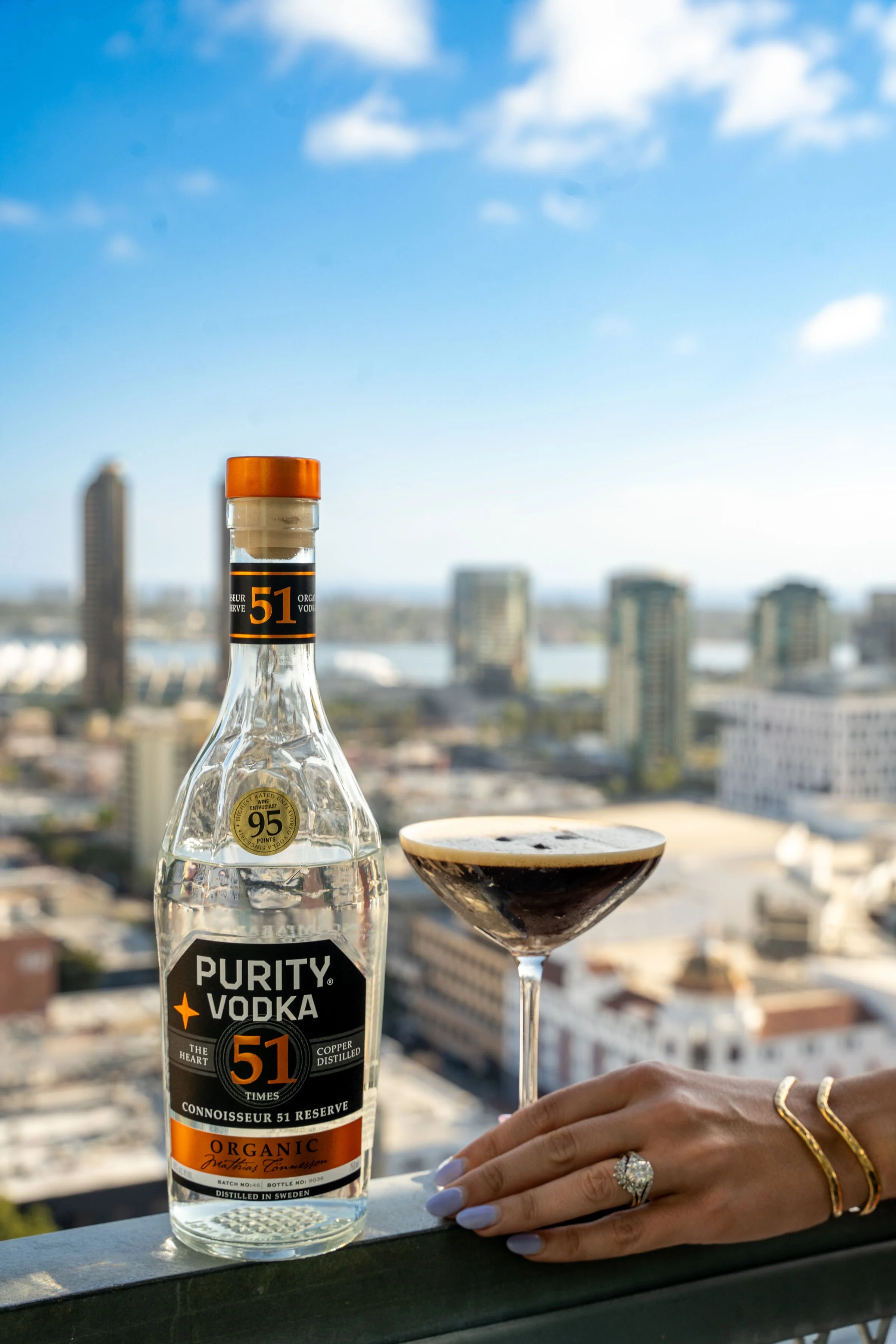A hand with jewelry resting on a railing next to a bottle of Purity Vodka and a cocktail glass with a dark drink, set against a cityscape with buildings and a blue sky with clouds.