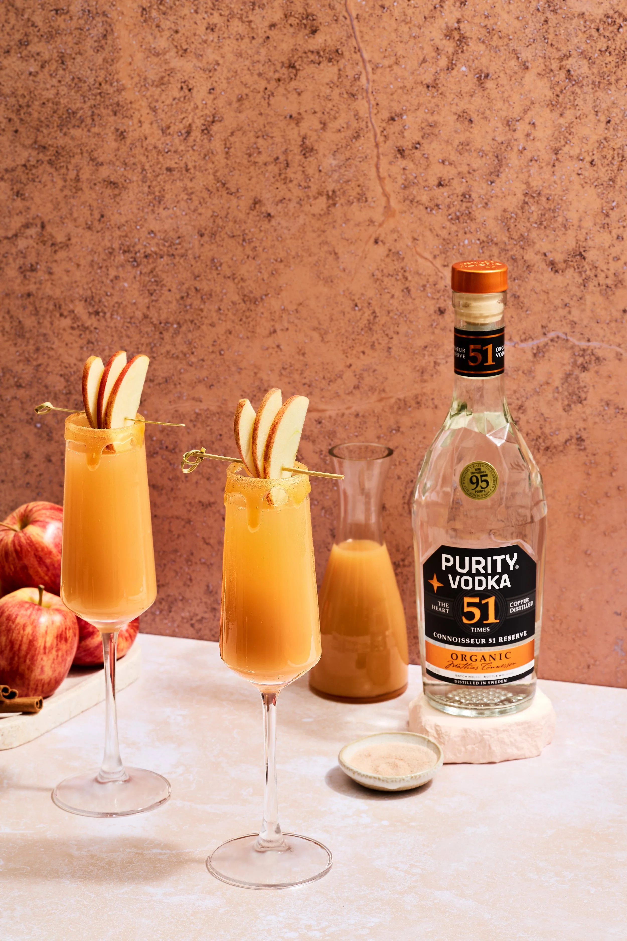 Two champagne flutes filled with apple cider garnished with apple slices, a bottle of Purity Vodka, a small bowl of sugar, apples, a small pitcher of apple cider, and cinnamon sticks on a light-colored surface with a textured pinkish background.