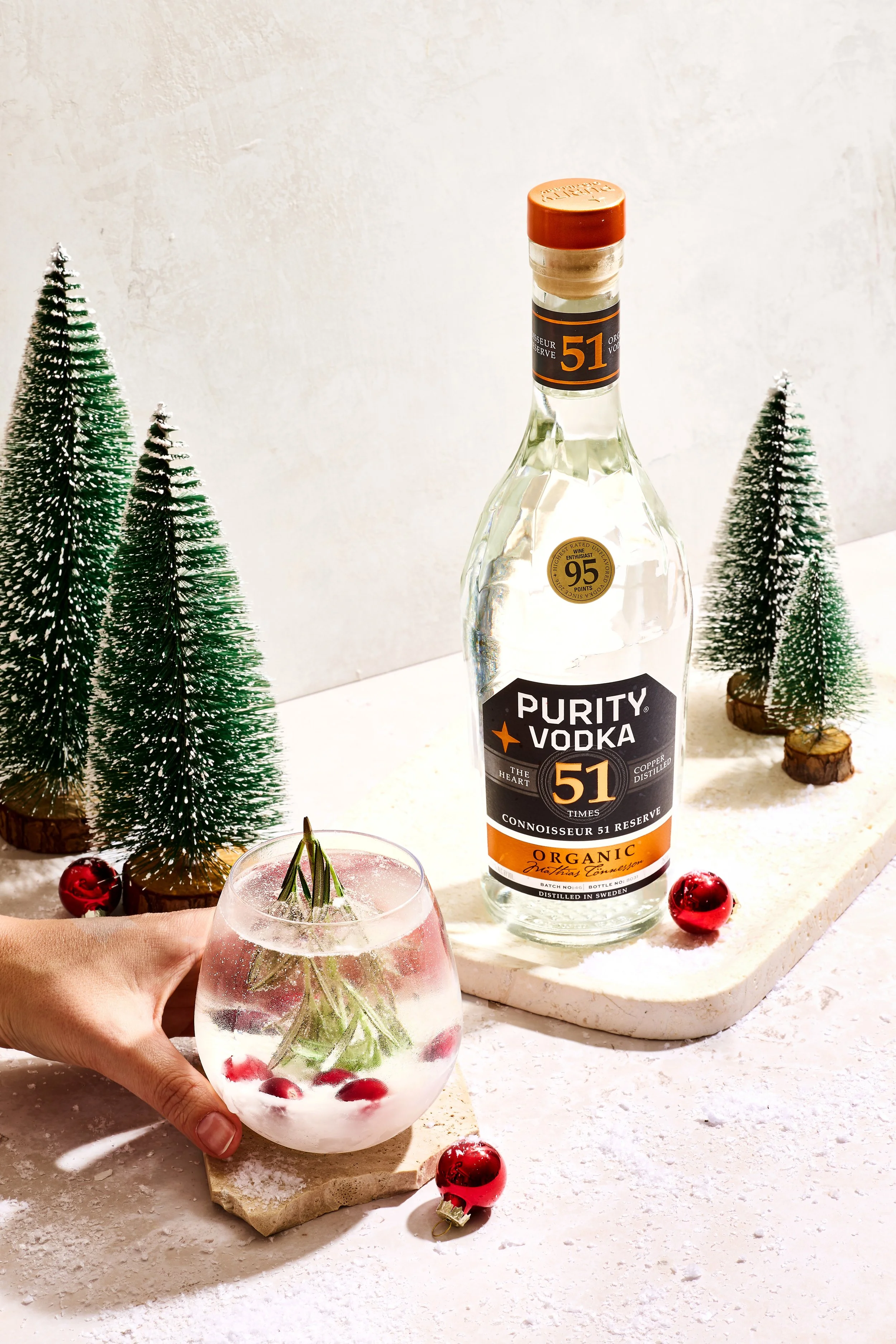 A bottle of Purity Vodka placed on a white surface with decorative miniature Christmas trees and red ornaments, a hand holding a glass of vodka infused with herbs and berries, and a marble serving platter.