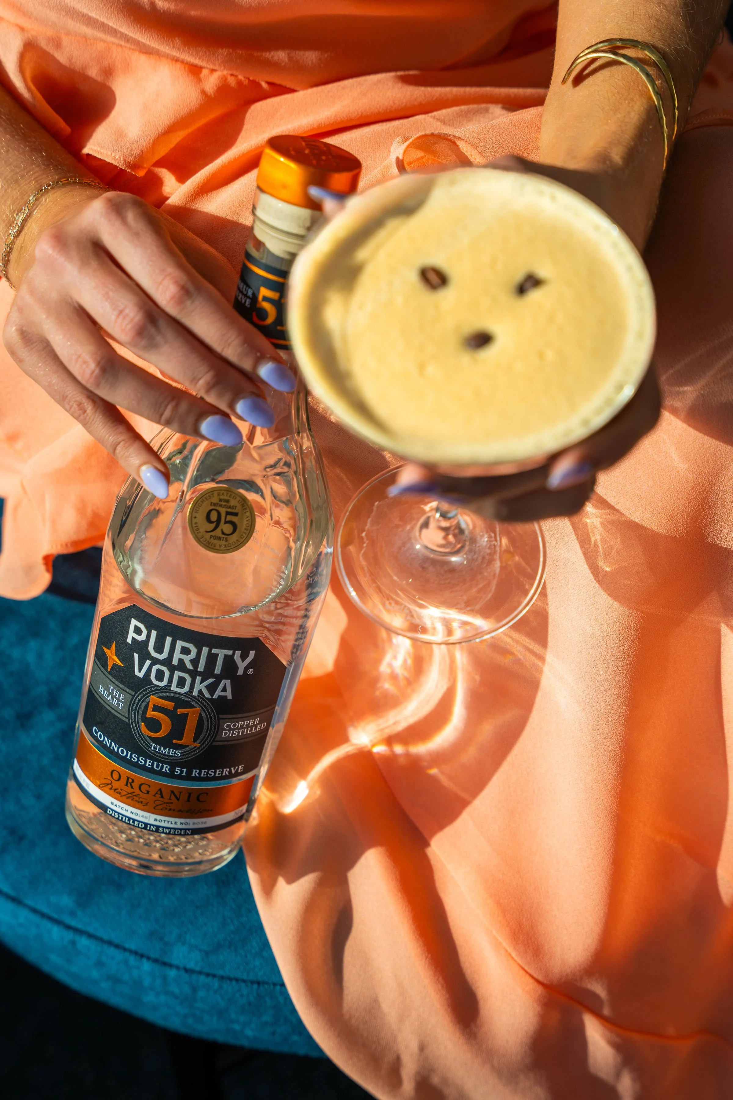 A person wearing gold jewelry holding a martini glass with a creamy beverage, garnished with coffee beans, and a bottle of Purity Vodka on a peach-colored fabric surface.