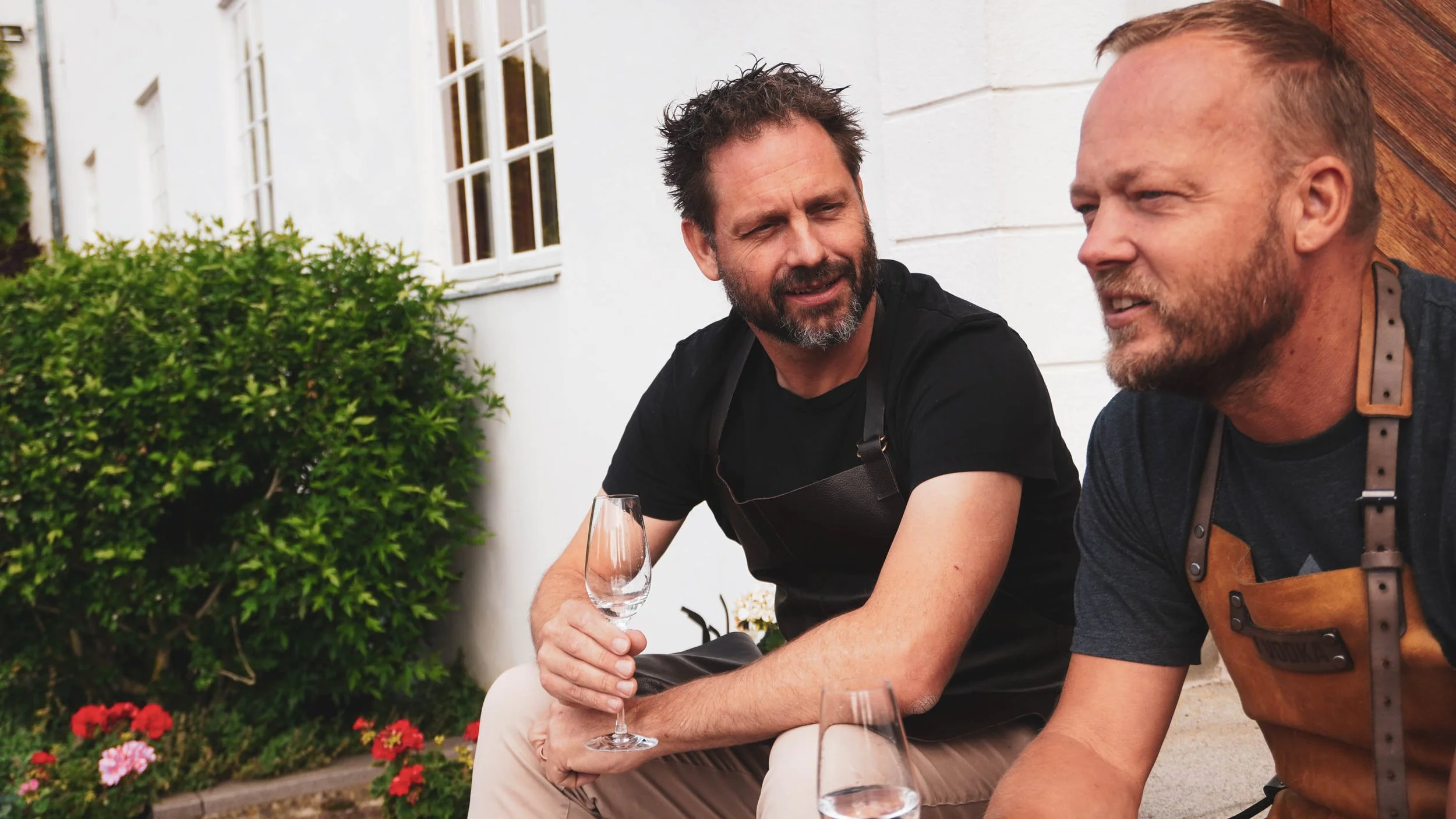 Two men sitting outdoors in conversation, holding glasses of wine, near a house with a white wall and windows, with bushes and flowers nearby.