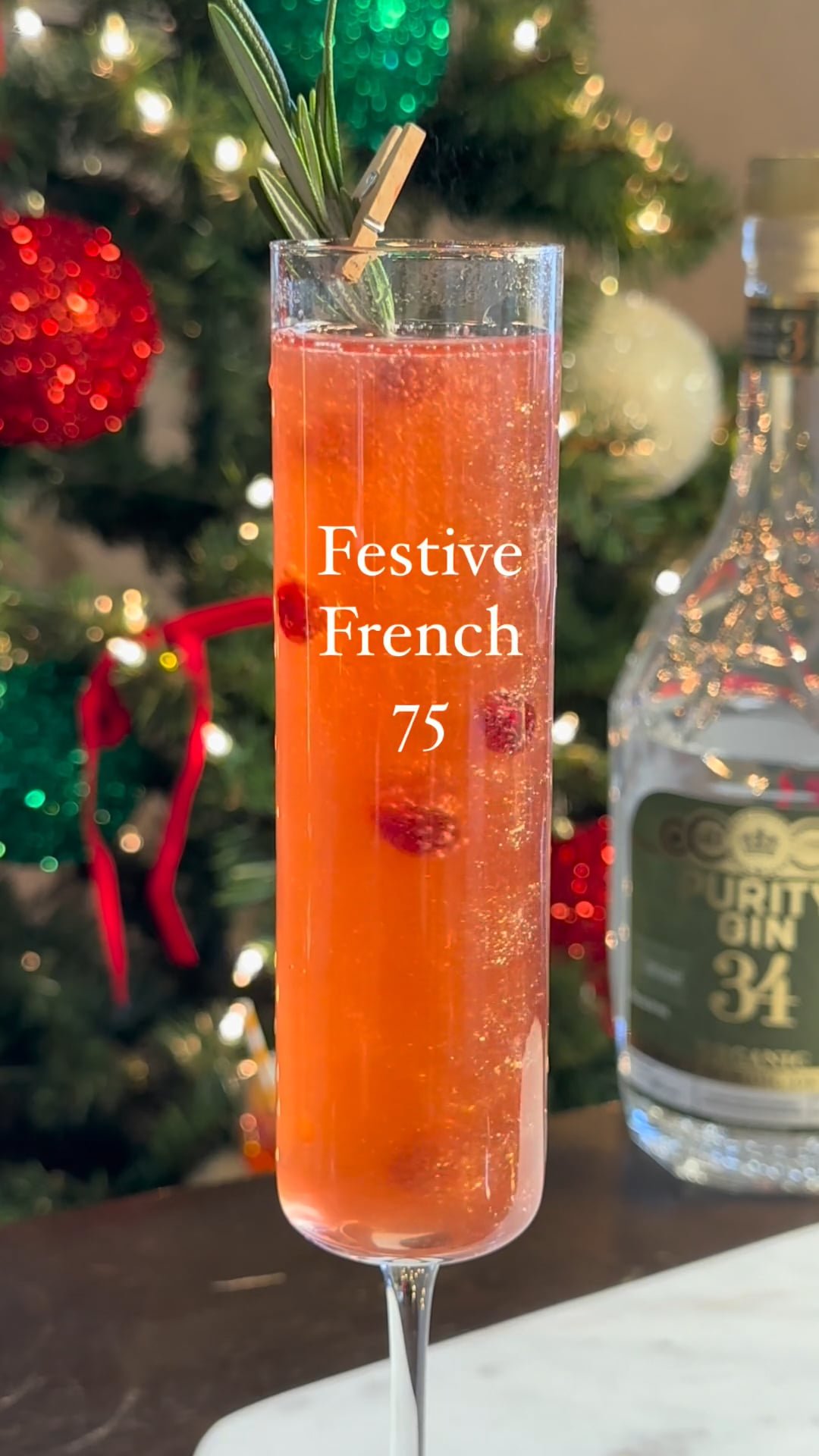 A tall glass of pink cocktail with berries and a sprig of rosemary, labeled 'Festive French 75', placed in front of a decorated Christmas tree.
