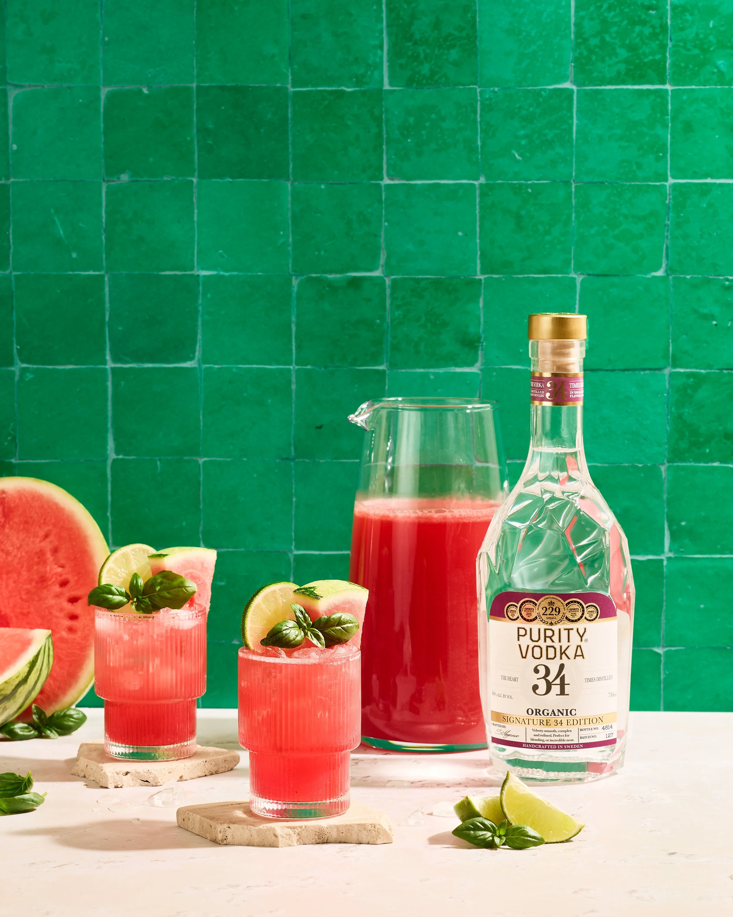 Close-up of a bottle of Purity Vodka in front of a pink drink with lime and basil garnishes in glasses, with a sliced watermelon, lemon, basil leaves, and lime slices on a white surface against a green tiled wall.
