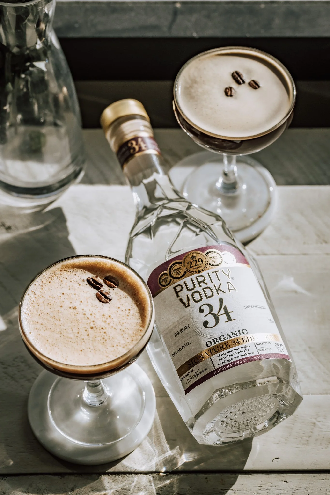 A bottle of Purity Vodka and two cocktails with coffee beans garnishing the drinks, set on a rustic table.