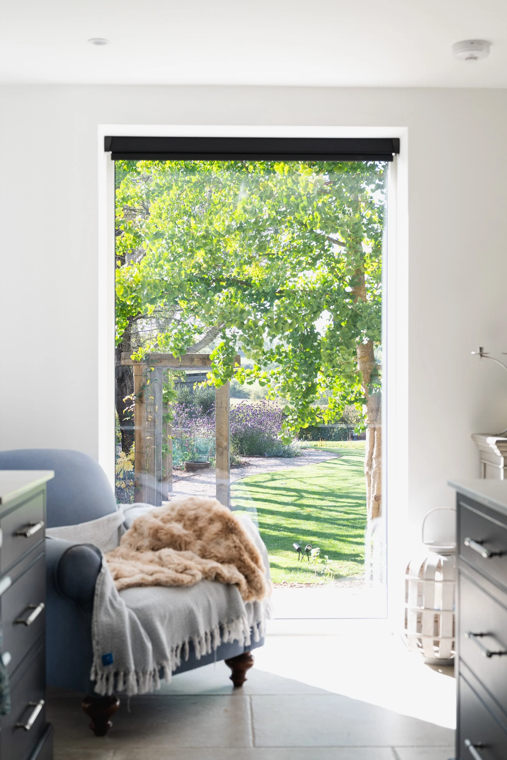 An armchair with a textured throw beside a tall window overlooking a lawn and trees.