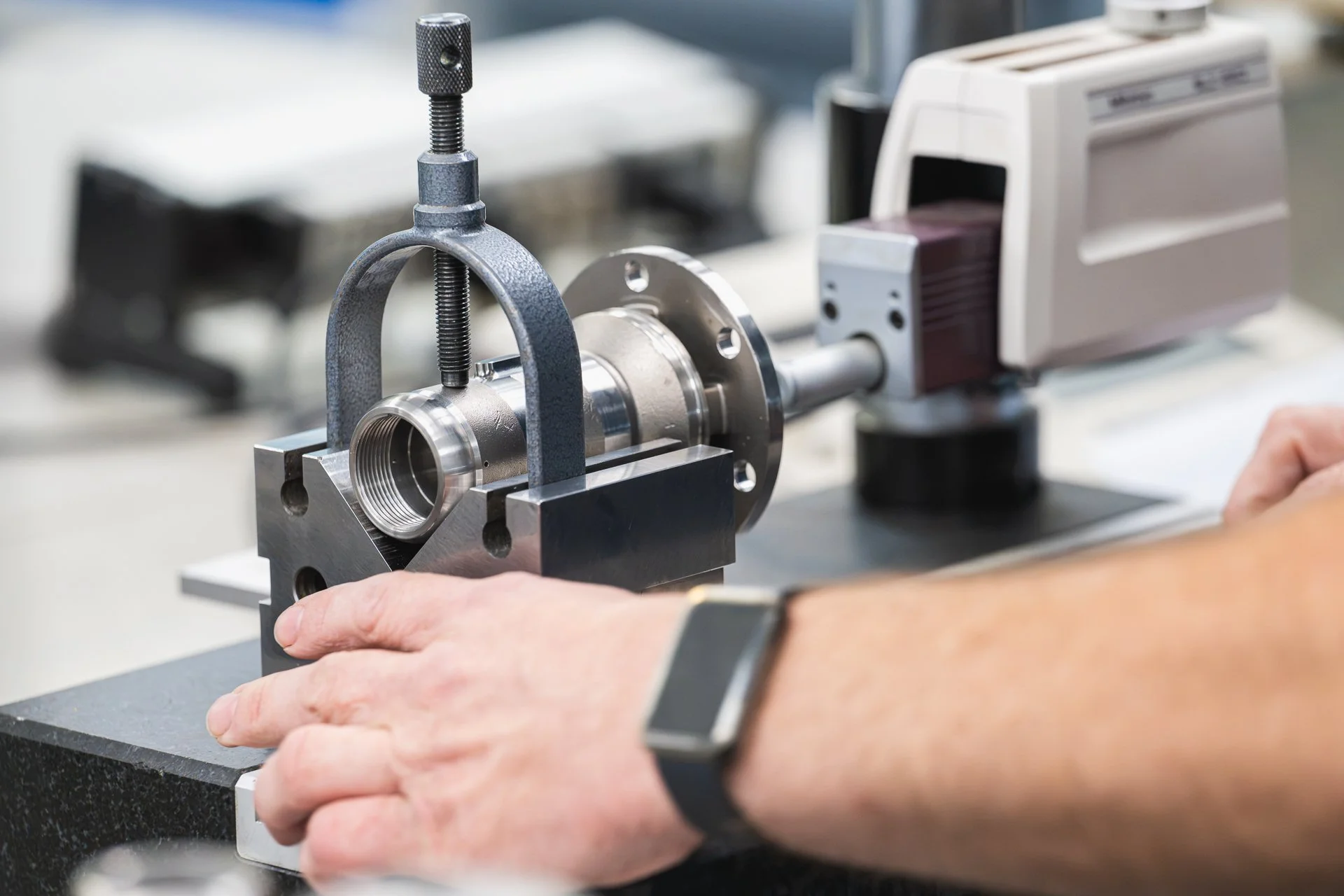 Hands positioning a machined metal component beneath measuring equipment.