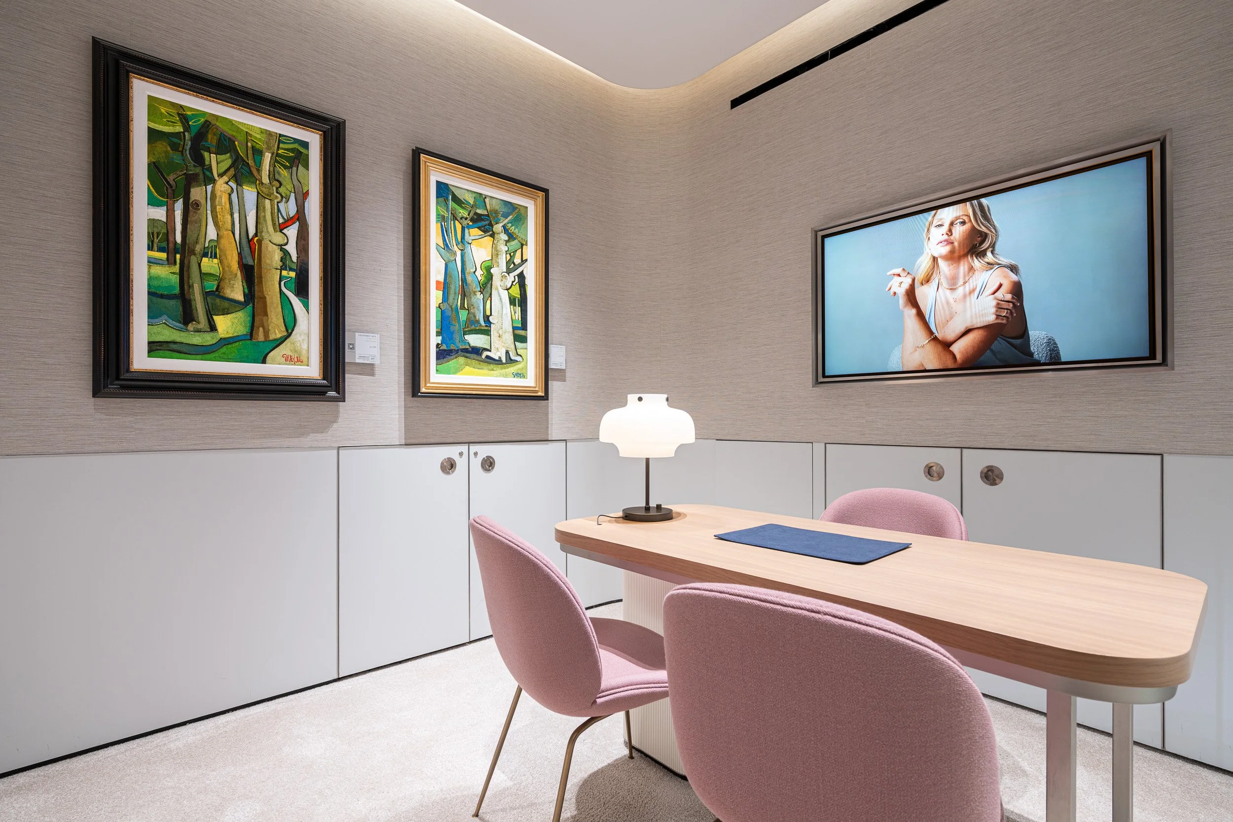 Two framed paintings beside a consultation table with pink chairs and a wall-mounted screen.