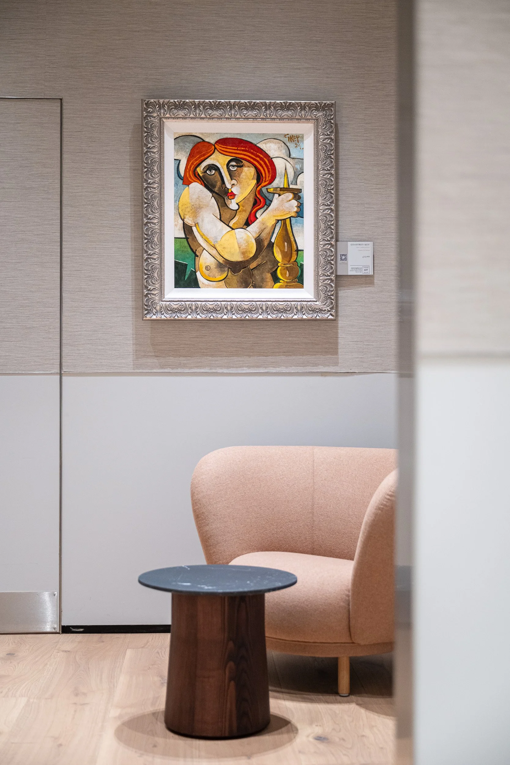 Framed painting of a head above a pink chair and dark side table.