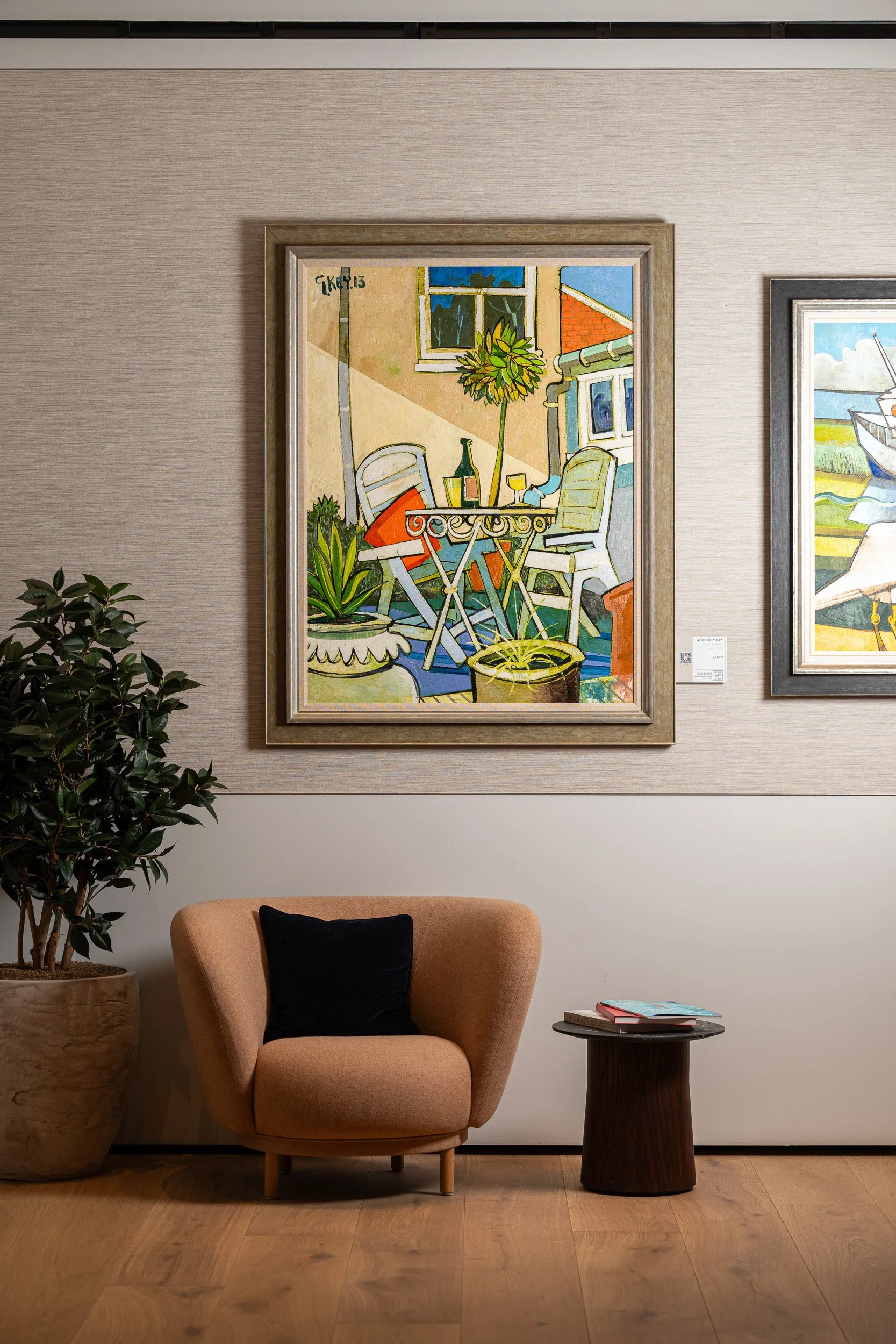 Large colourful framed painting above a pink chair and small side table.
