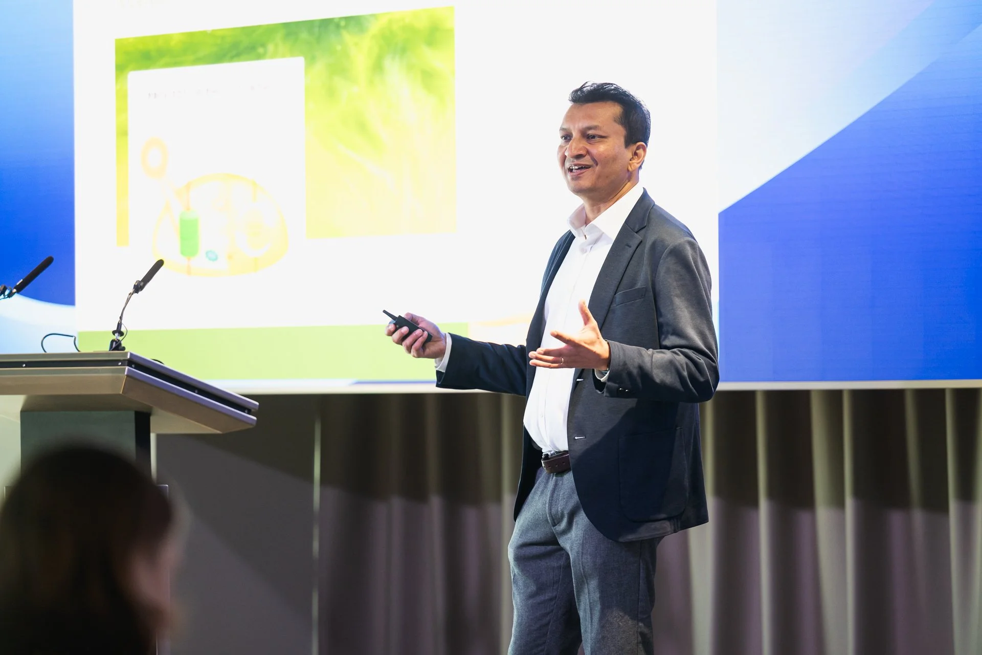 A man in a dark suit gesturing beside a lectern, with a presentation displayed behind him.