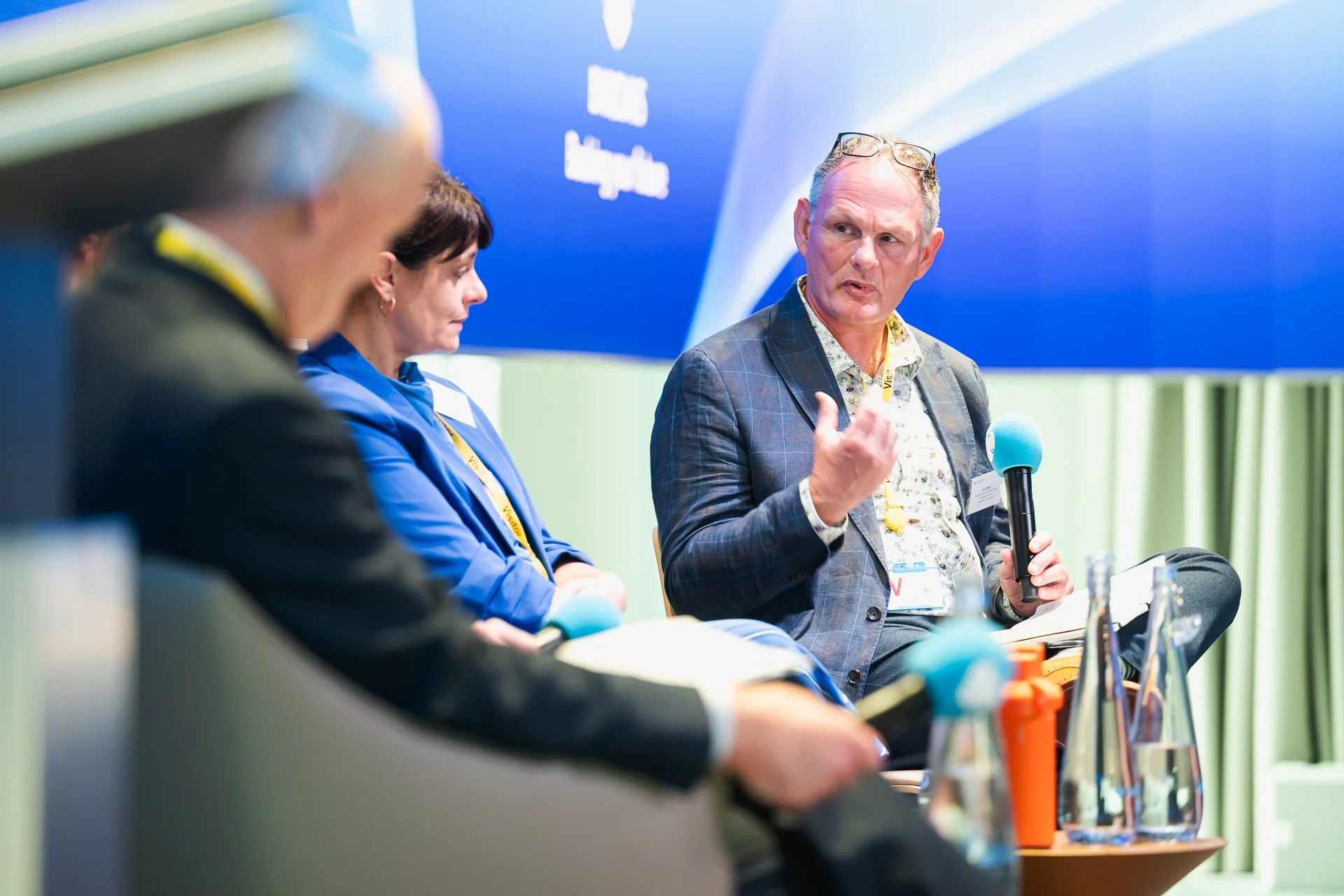 A seated panellist holding a microphone and gesturing, with other panellists beside him.