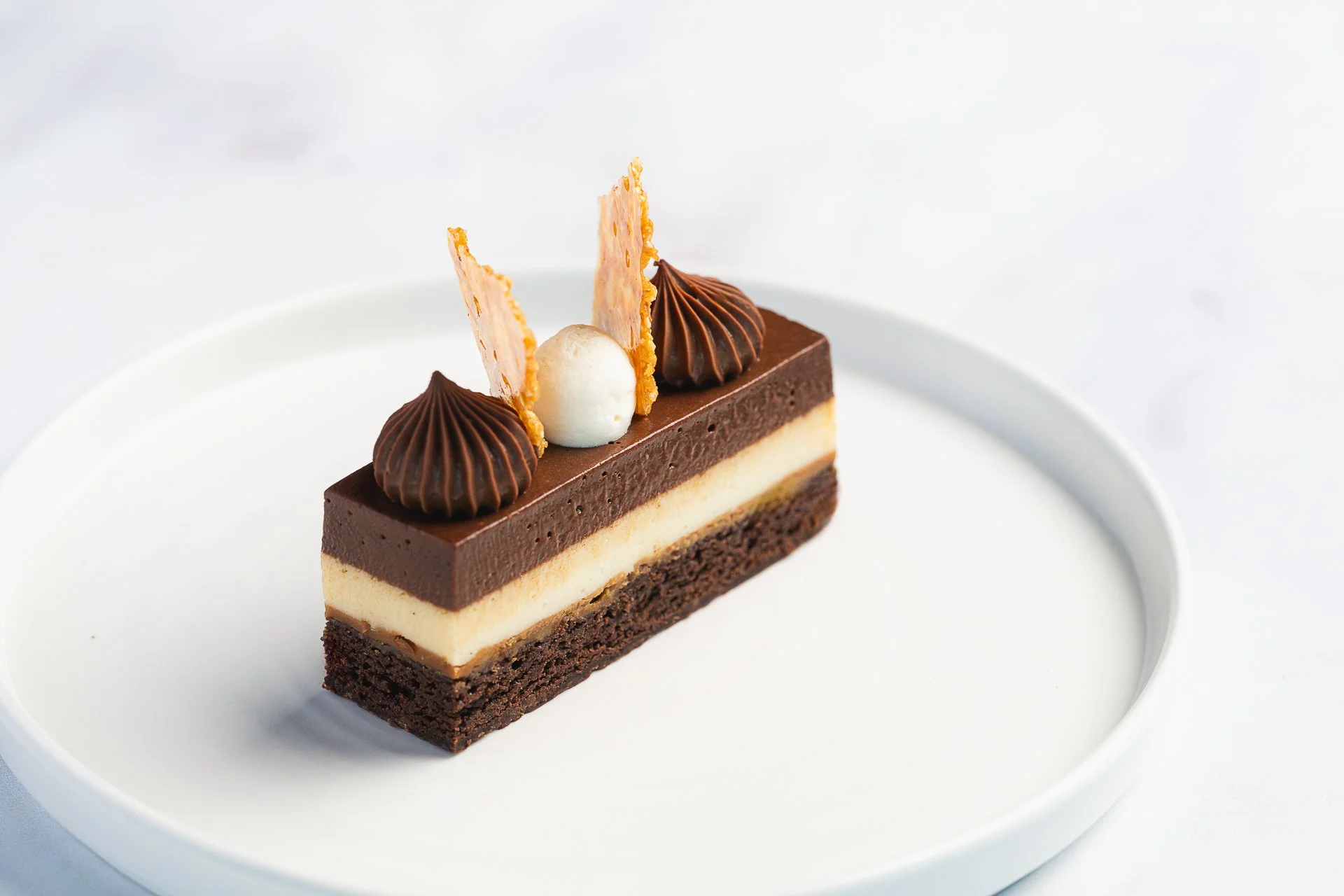 A rectangular layered dessert topped with piped chocolate and thin decorations on a white plate.