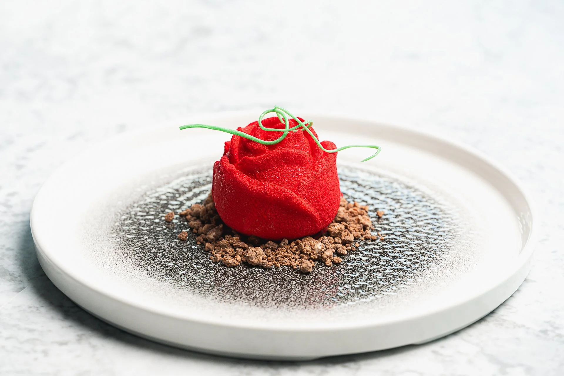A red rose-shaped dessert with a green decoration on a white plate.