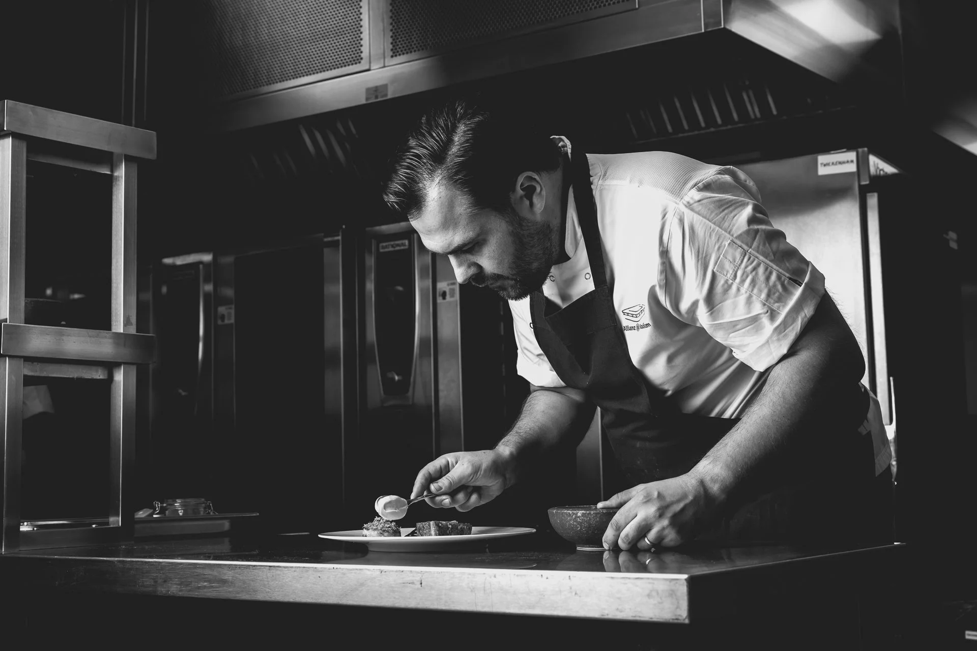 Black-and-white photograph of a chef bending over a plate and adding a finishing detail with a spoon.