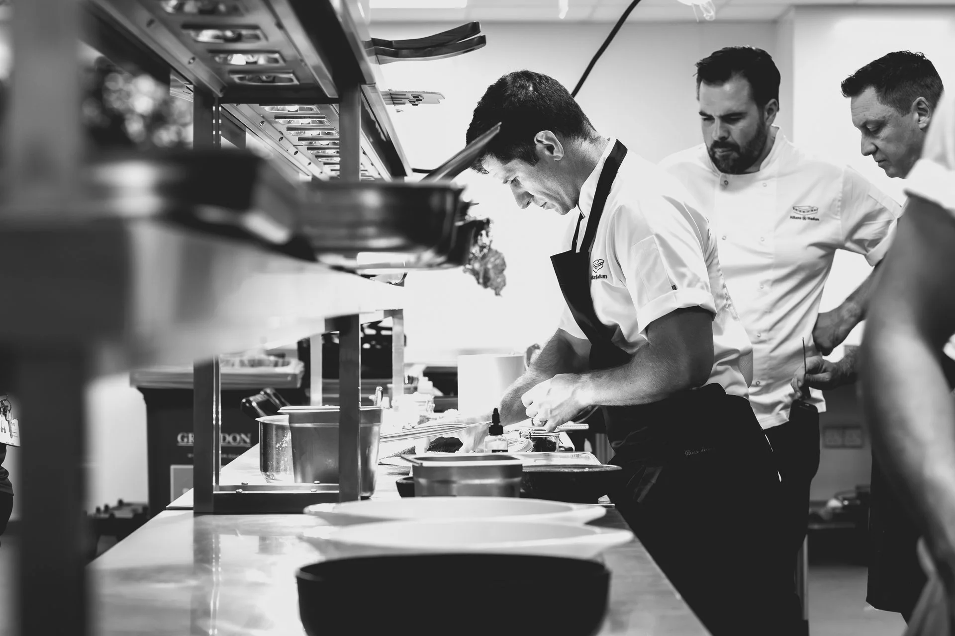 Black-and-white photograph of a chef leaning over a plate with other chefs beside the kitchen pass.